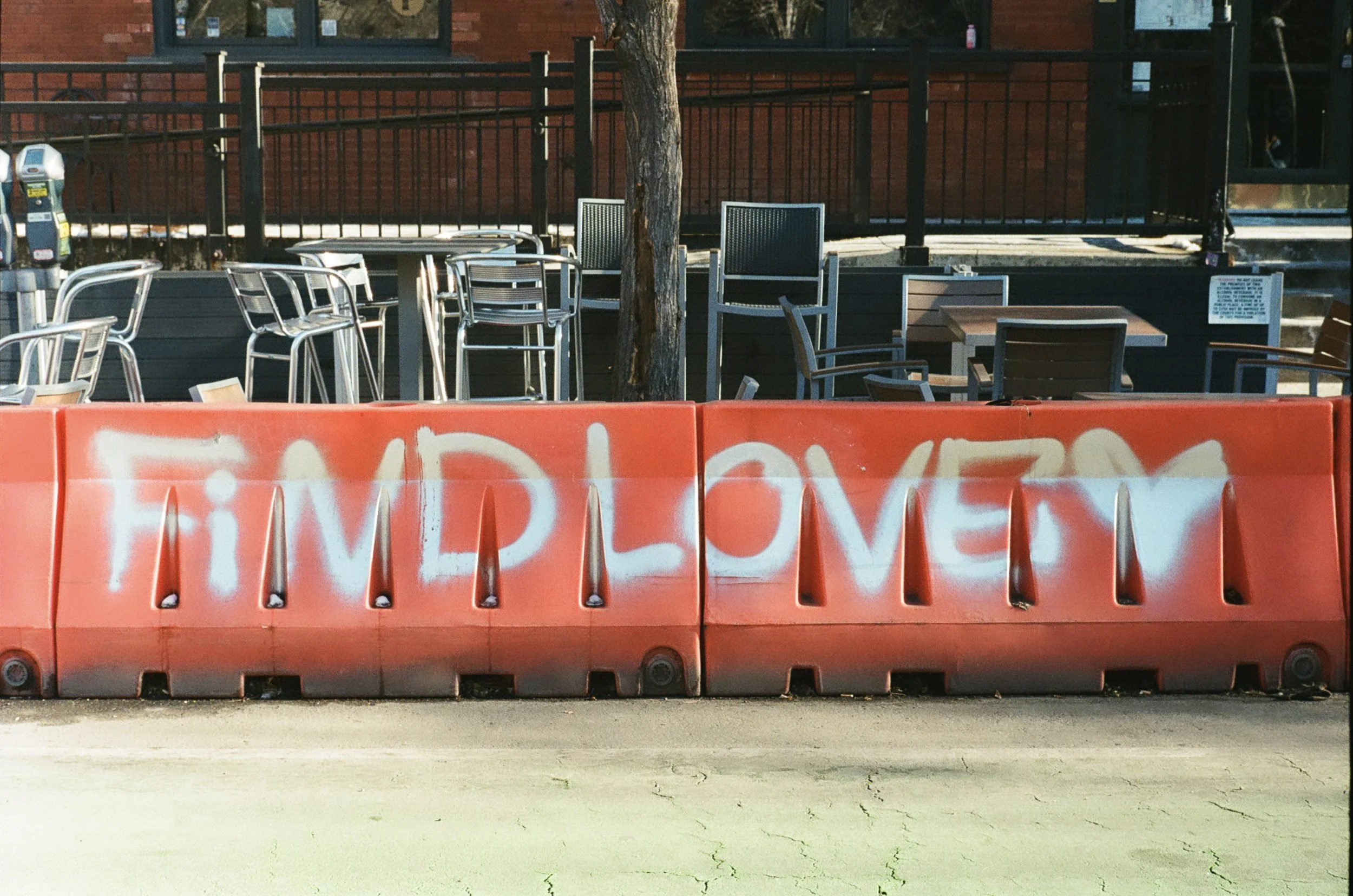 A row of outdoor chairs and tables behind a red barrier with graffiti, with a tree and a brick building in the background.