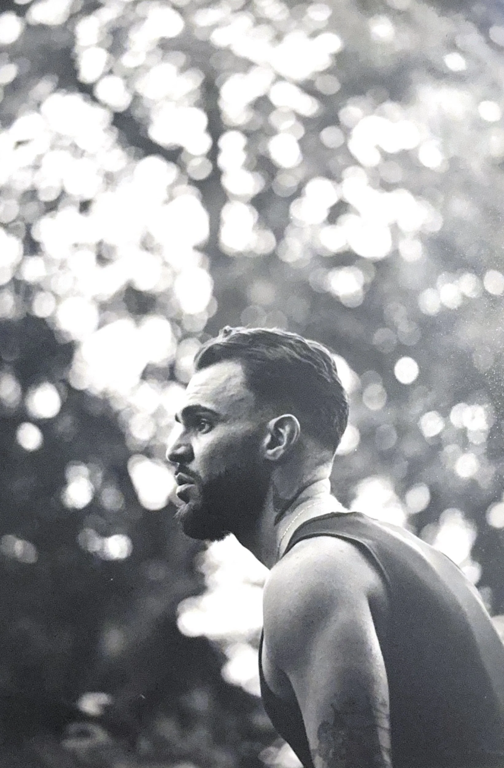 A black and white photograph of a man in a sleeveless shirt with a muscular build, focused and looking to the side, with a background of shimmering light effects.