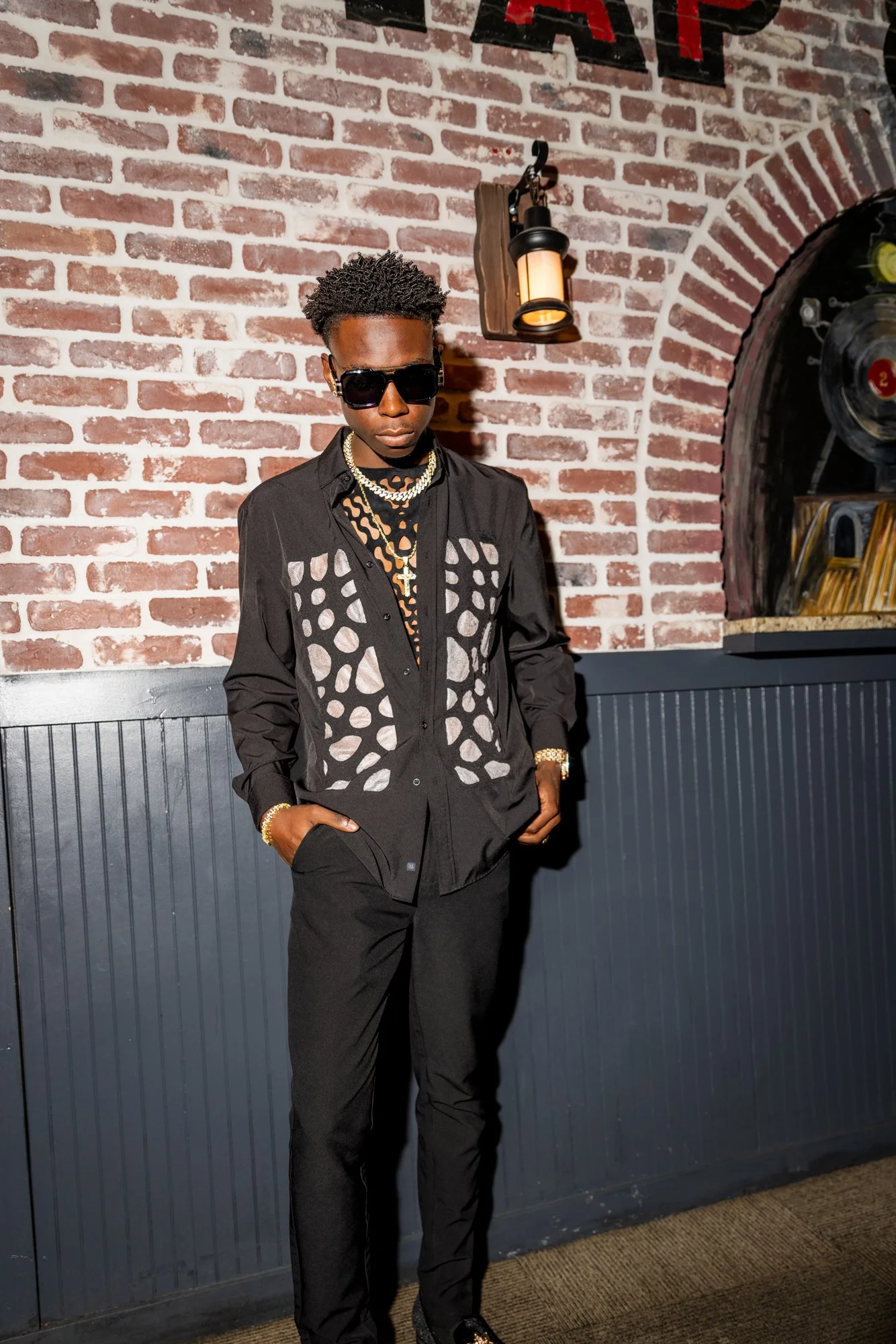 A young man dressed in black with patterned shirt, wearing sunglasses, gold chains, and bracelets, standing against a brick wall with some visible artwork or signage. He has one hand in his pocket and is looking downward.