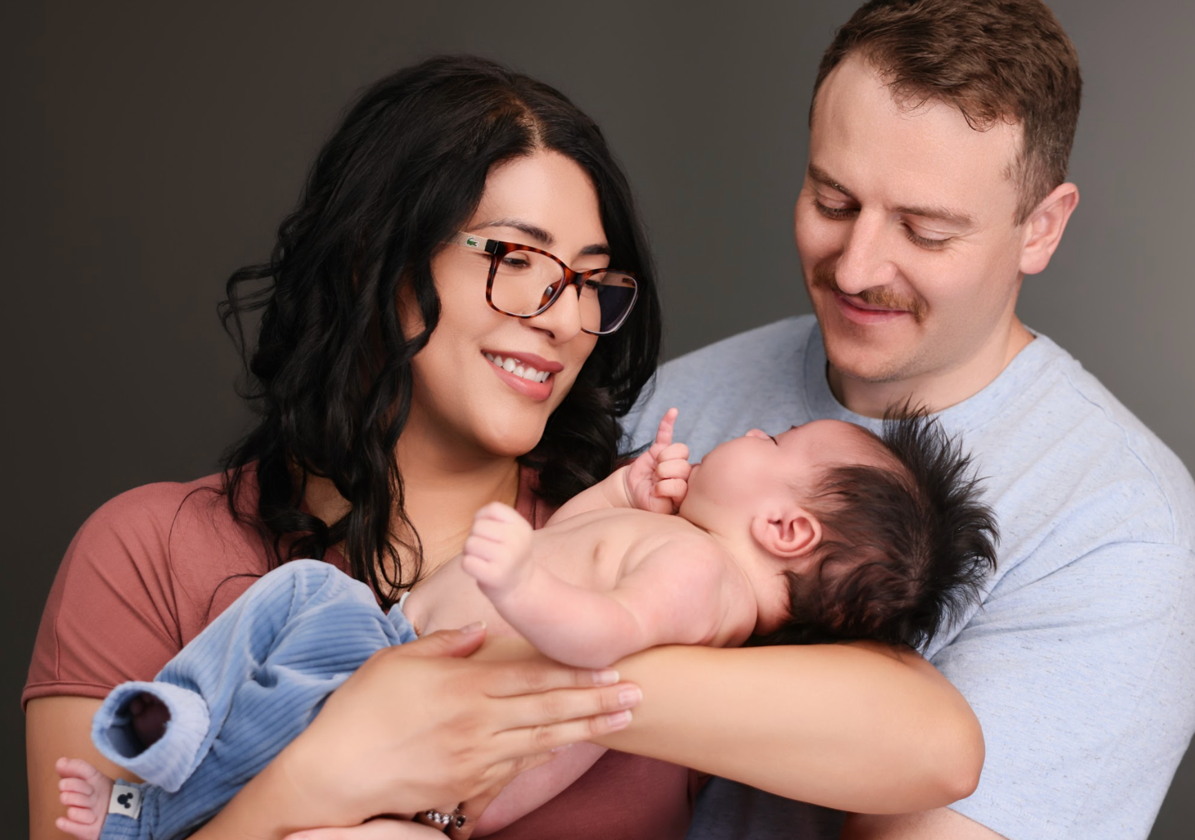 A smiling woman with glasses and dark hair is holding a newborn baby. A man is smiling at the baby, cradling him in his arms. All three are looking at each other affectionately against a dark background.