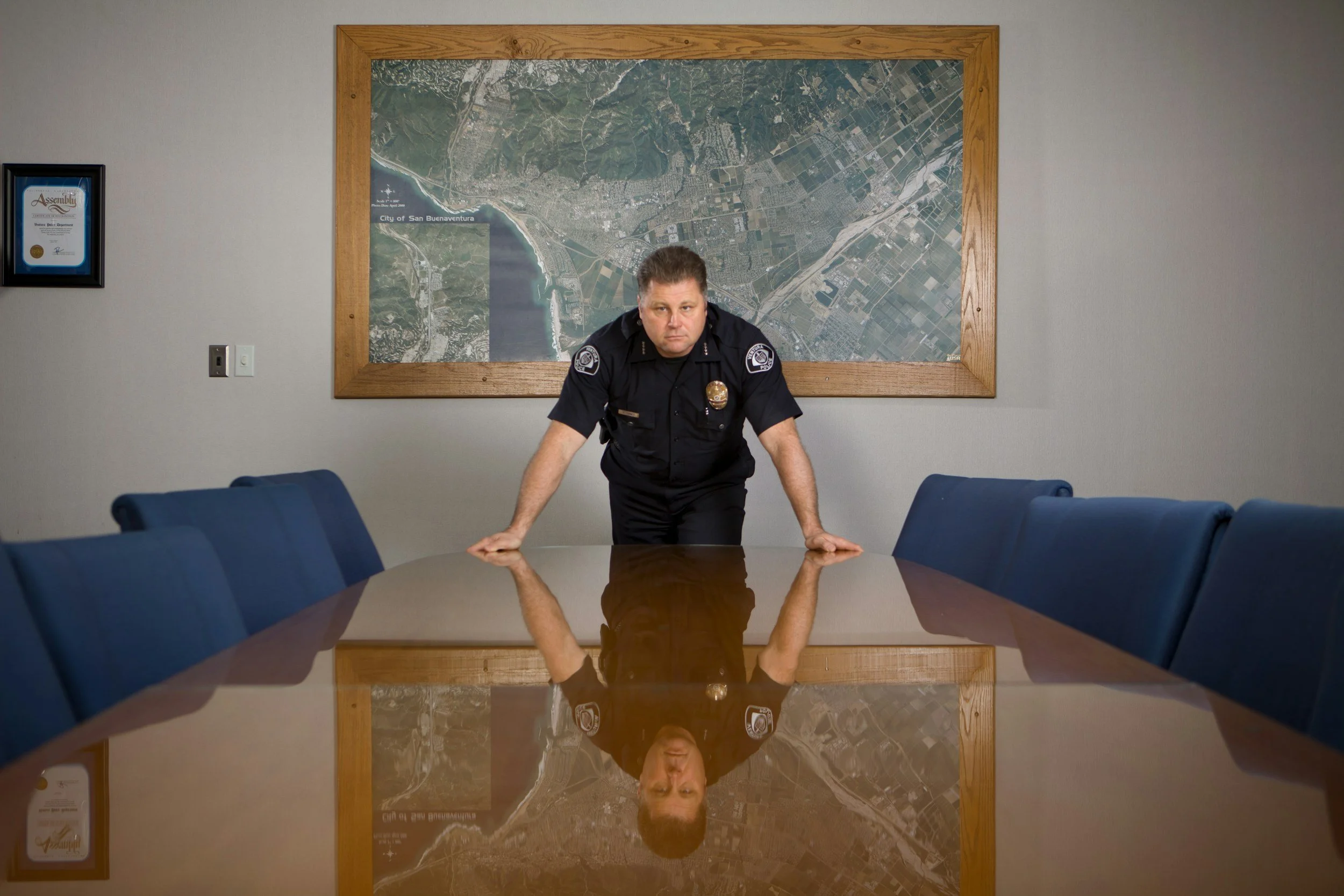 A police officer leaning on a conference table in a room with a large map of San Buenaventura on the wall behind him. A framed certificate is on the left wall of the room.