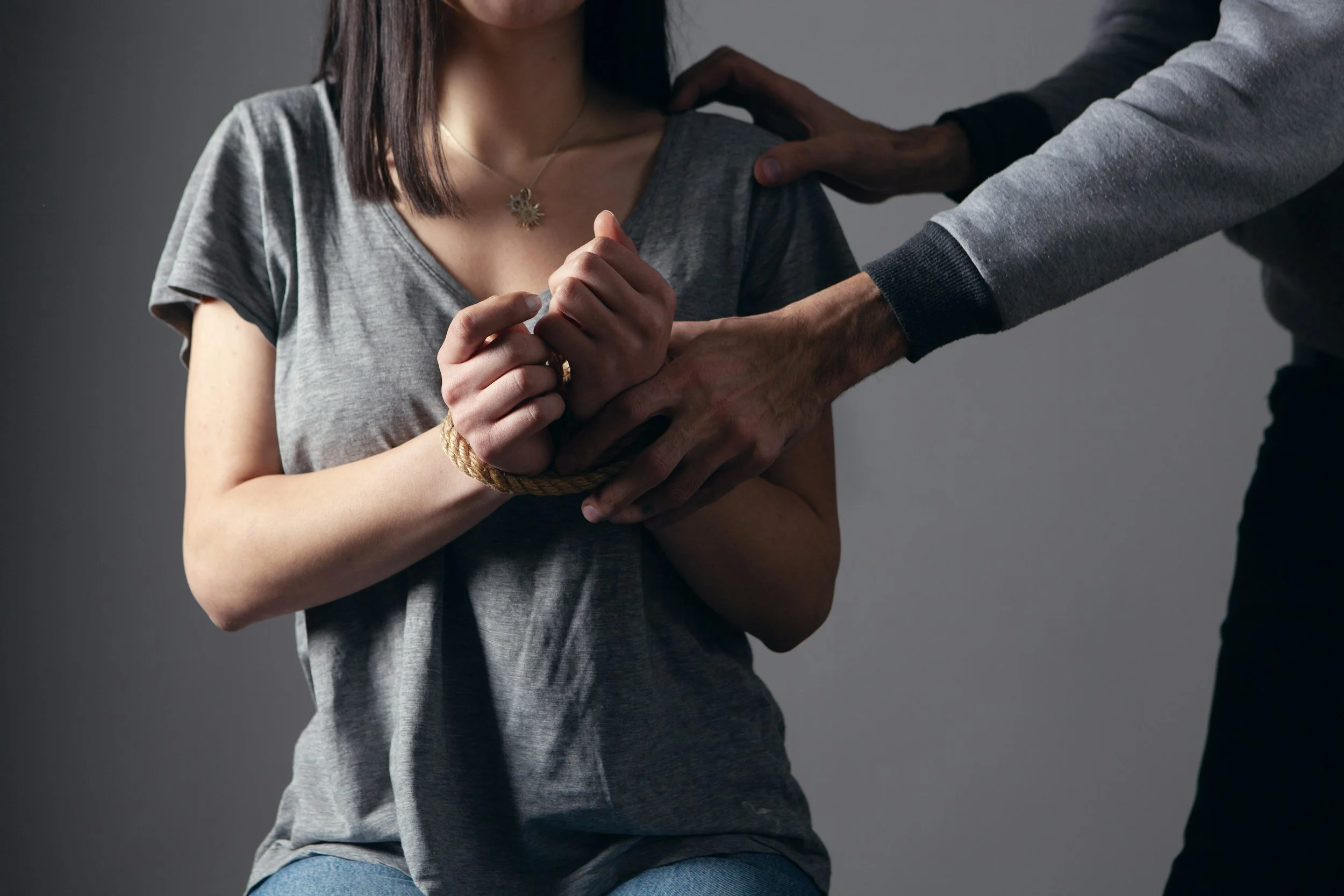 A woman with medium-length brown hair wearing a grey t-shirt and necklace is being restrained by a man with dark skin, wearing a grey and black long sleeve shirt. The man is holding her wrist and placing his hand on her shoulder, which is bound with a rope.