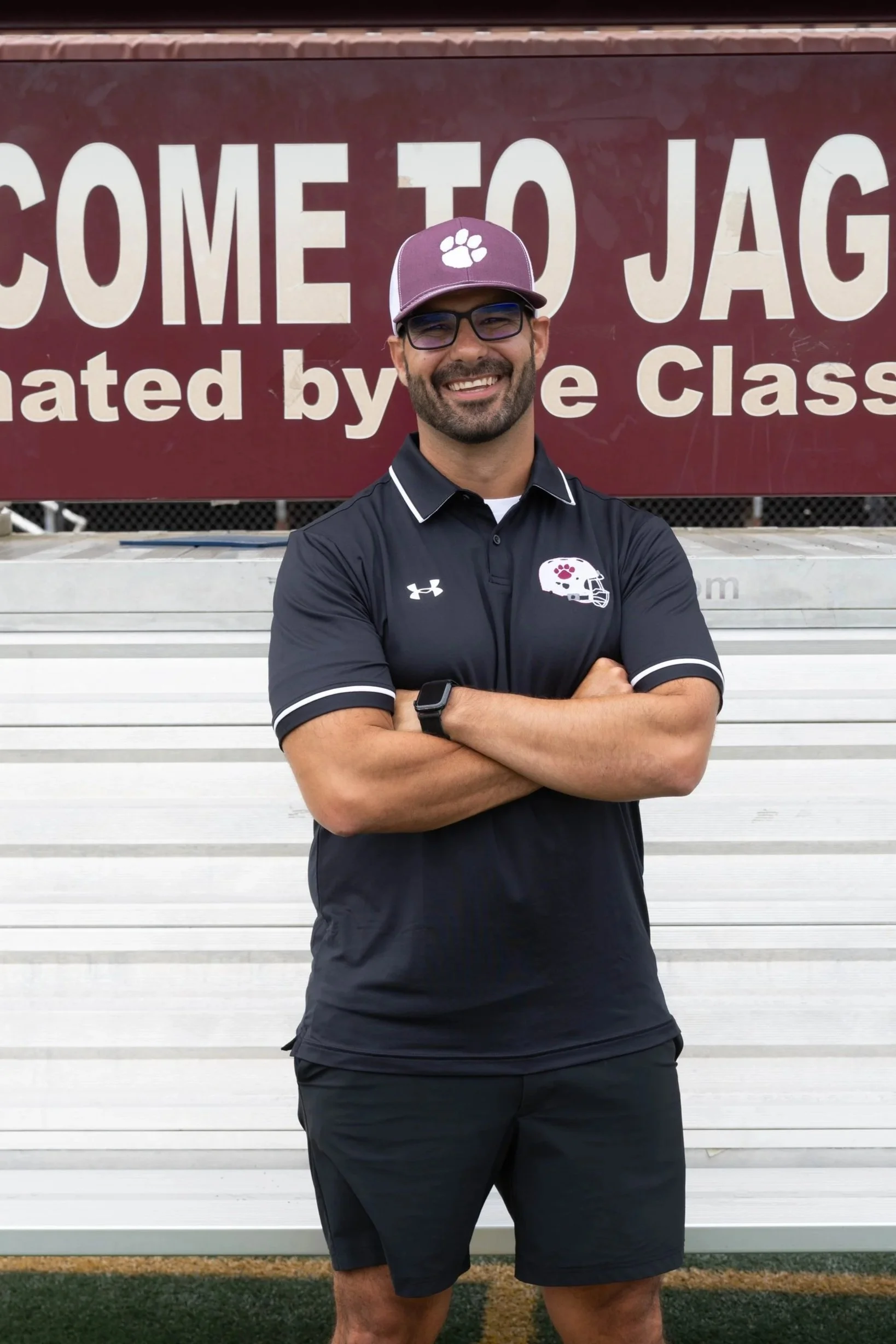 A man in a burgundy polo shirt with G V Football logo stands on a running track with arms crossed, smiling, in front of a sign that reads 'ONE NESS' with a football-shaped design on a landscaped hill.
