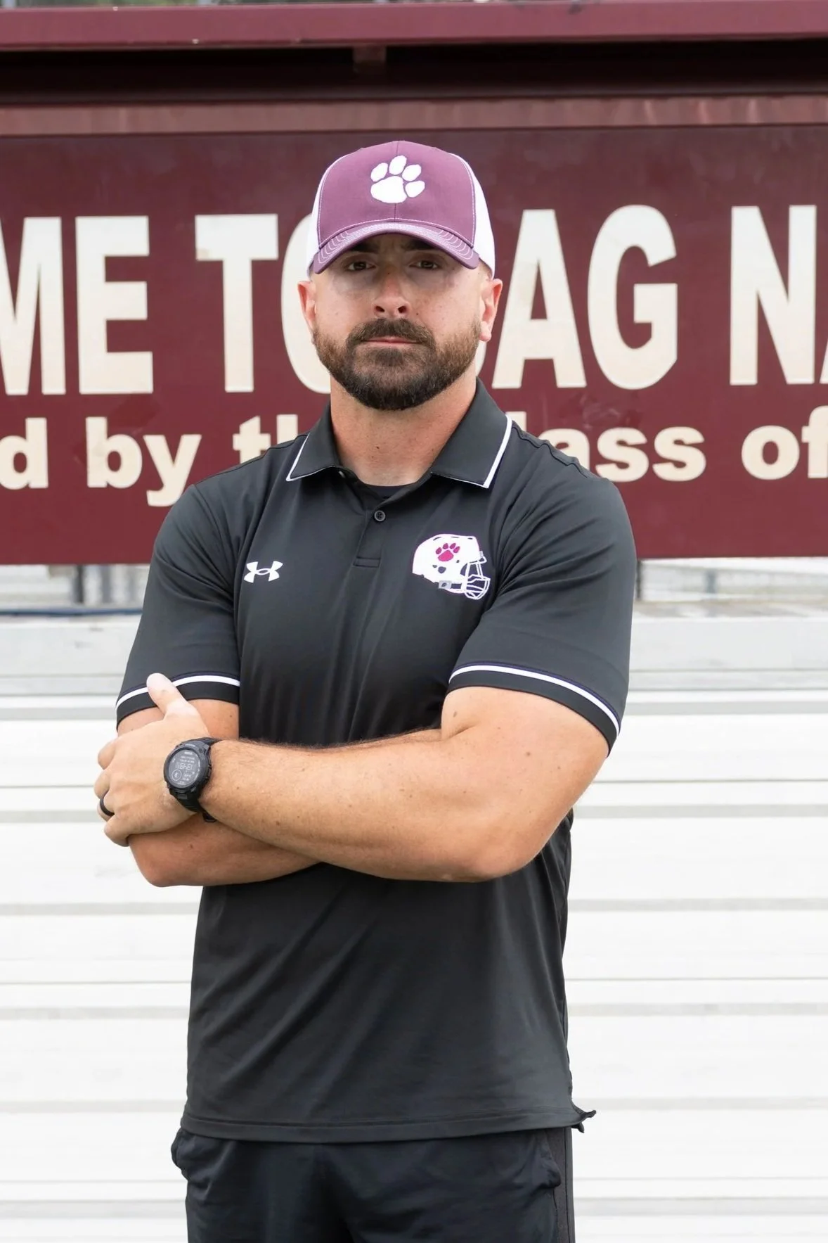 A man standing on a red running track with arms crossed, wearing a maroon golf shirt with a logo, black shorts, and a gray and black baseball cap, with a rock garden and a sign that says 'ON TRACKS' in the background.