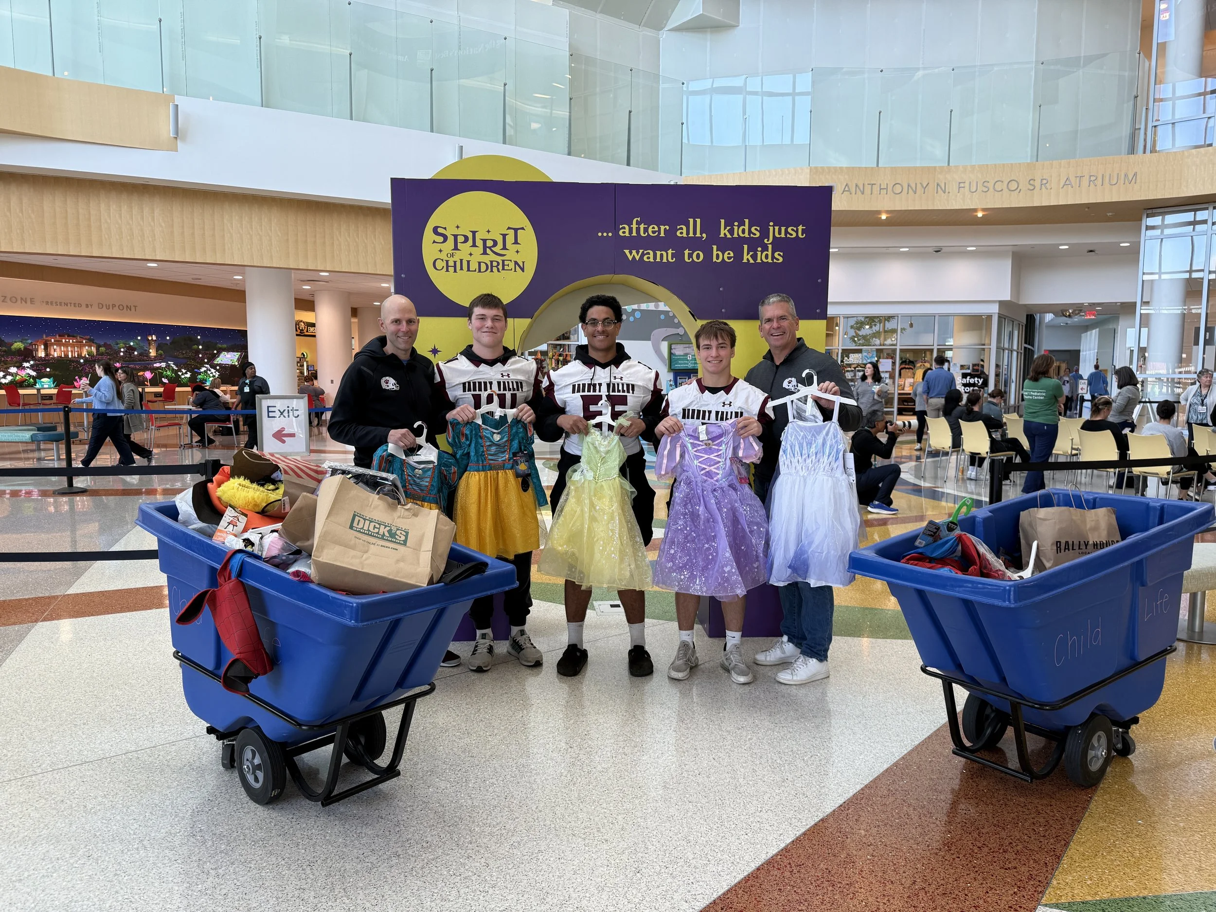 A group of five people, including children in sports jerseys, standing inside a shopping mall, holding dresses, with shopping bags and carts nearby, in front of a sign that reads 'Spirit of Children' and 'after all, kids just want to be kids'.