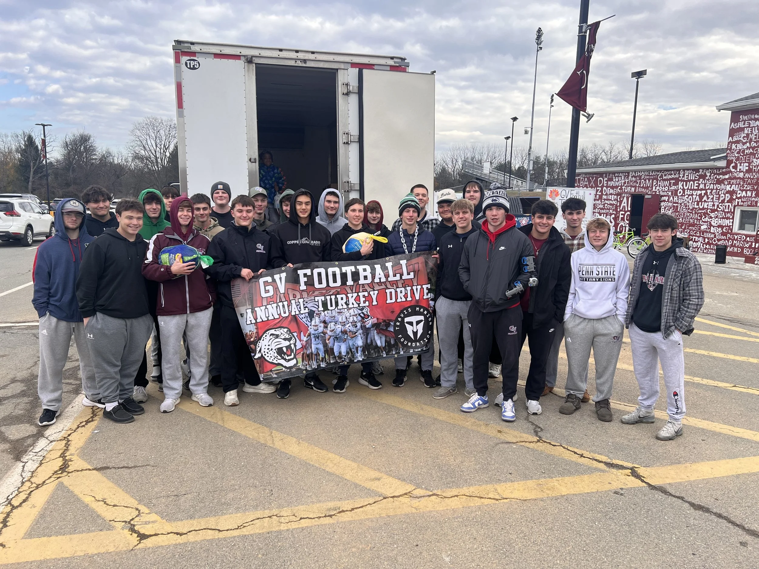 A group of young boys and teenagers standing outdoors in a parking lot, holding a banner that reads 'GV Football and Turkey Drive.' They are dressed in casual and athletic clothing, with some wearing hoodies and jackets. There is a large white truck behind them with its back open, and a boy is sitting inside the truck. The background has trees, parked cars, and a maroon building with white graffiti-style writing, along with flags and outdoor lights.