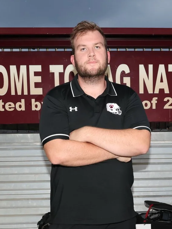 A young man in a maroon GV Football polo shirt and a cap stands on a running track, with a sign saying 'GREATNESS' behind him on a hill with rocks and plants.