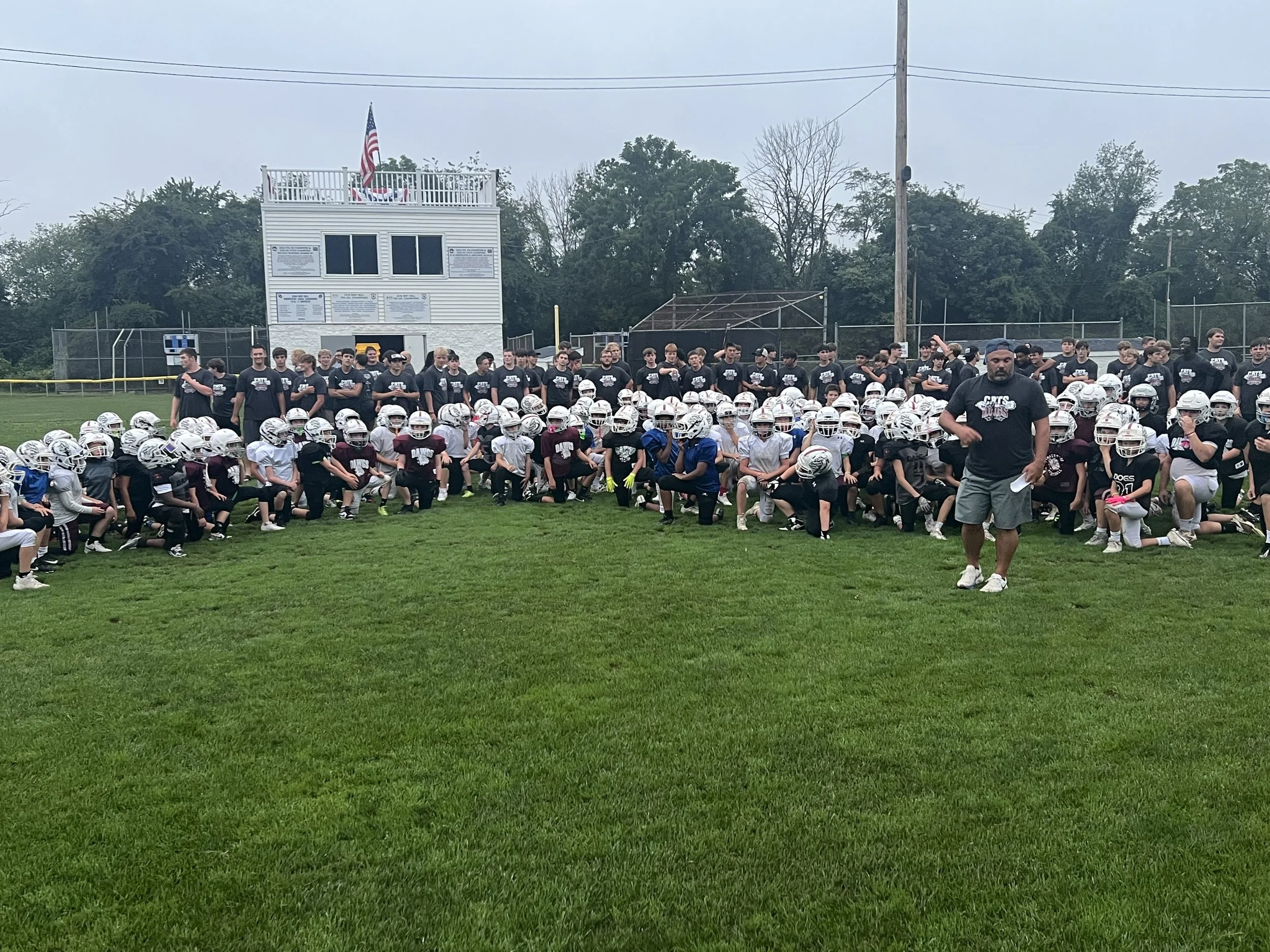 A large group of young football players sitting and standing on a sports field, wearing helmets and uniforms, with coaches and staff behind them. There is a white building in the background with an American flag on top, and trees around the field.
