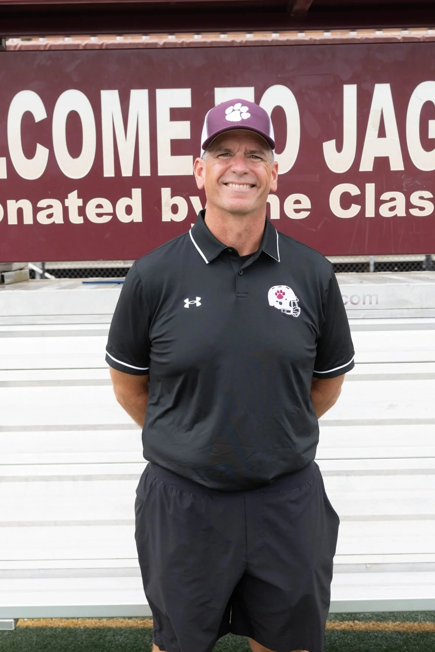 A man in a maroon polo shirt and a gray cap stands on a track field with arms crossed, smiling, with a football field emblem and landscaping in the background.