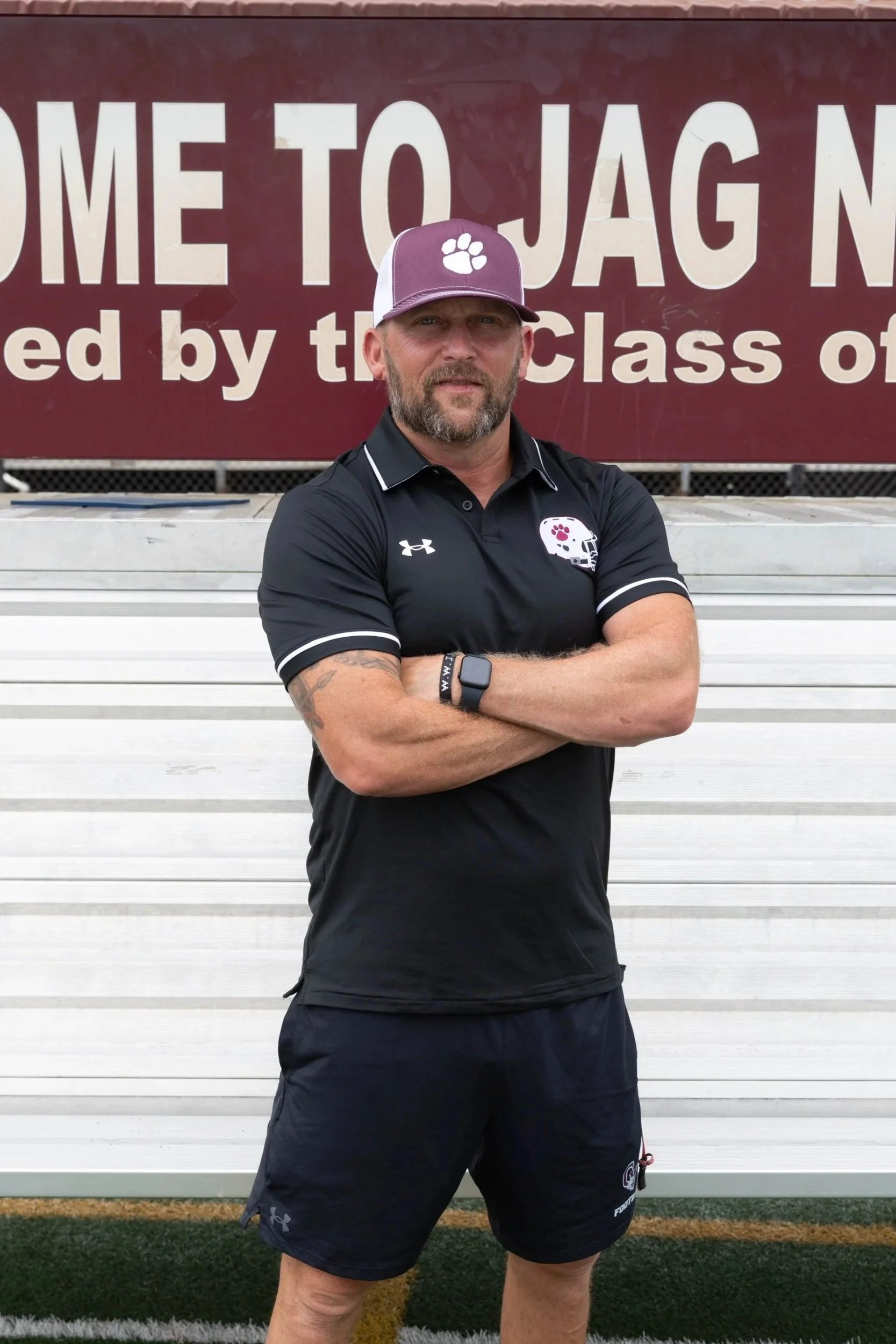 A man with a beard and mustache standing on a red running track, crossing his arms, wearing a maroon football shirt with a logo, a gray and black cap, with a sign that says 'ONENESS' in the background.