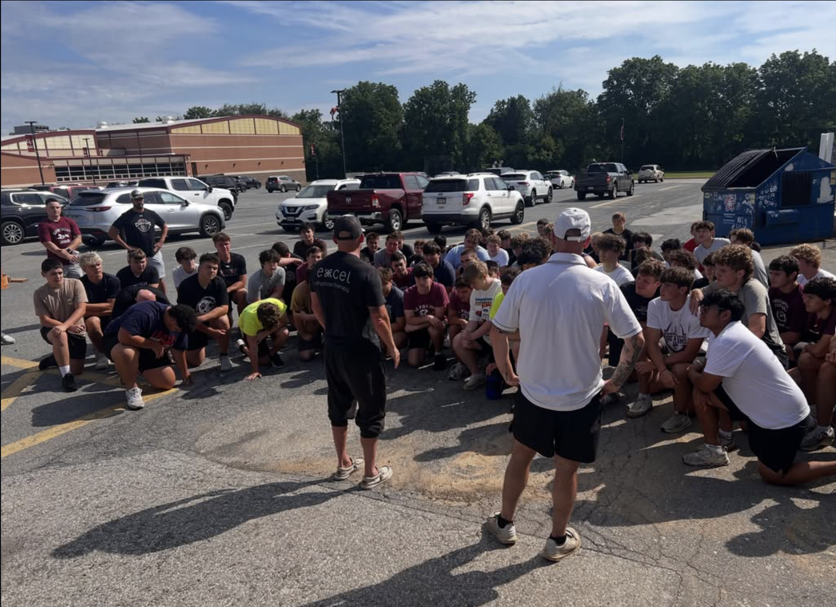 A large group of teenage boys sitting on the pavement in a parking lot listening to two men standing in front of them. The background shows parked cars, a blue dumpster, a building, and trees under a partly cloudy sky.