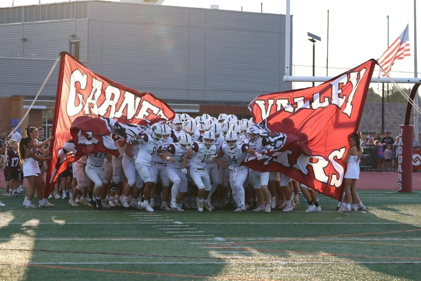 Football team in maroon uniforms with white helmets huddling behind a large banner reading 'GARNET VALLEY' on a sunny football field.