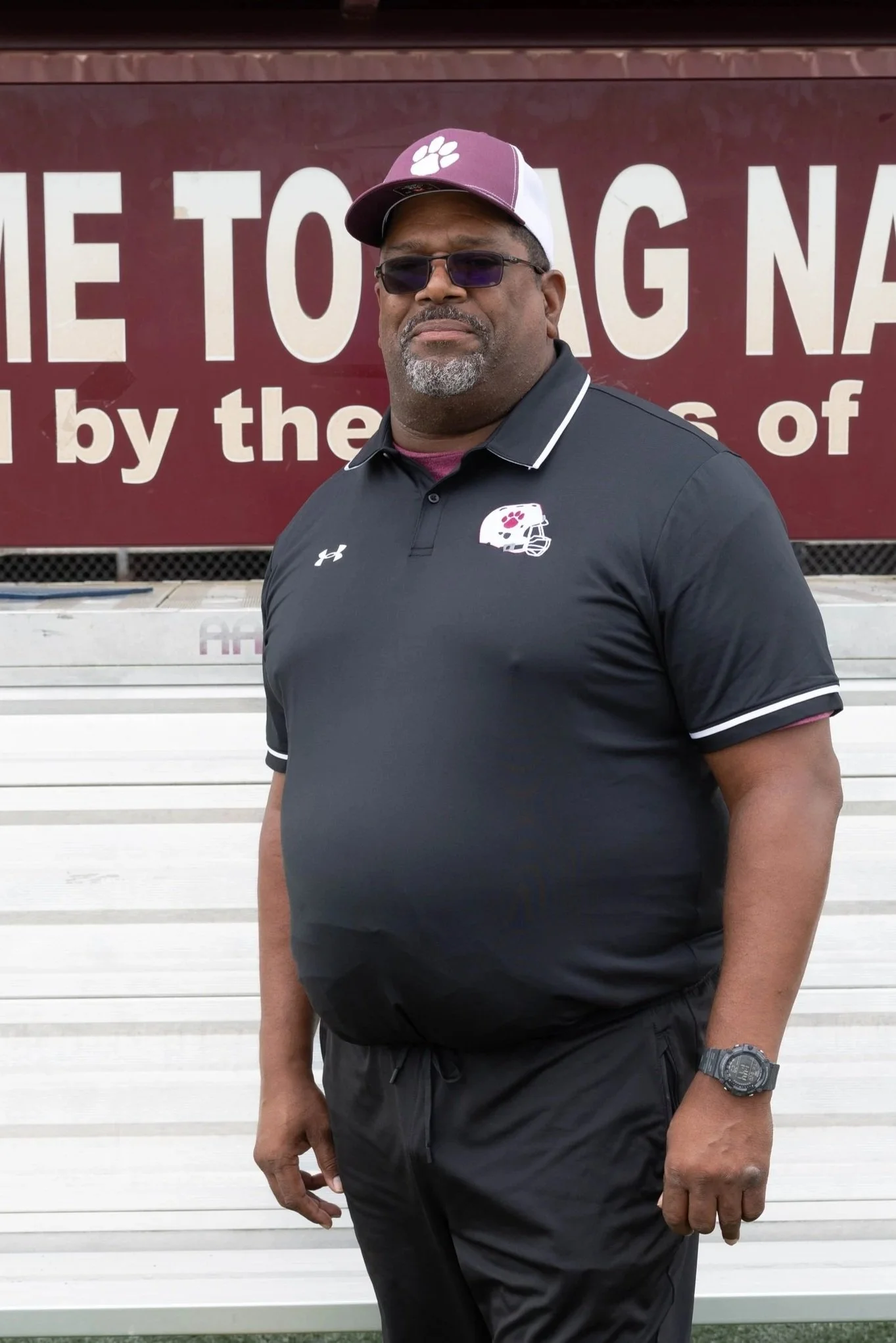 A man standing outdoors on a running track, wearing a maroon polo shirt, sunglasses, and a cap, with a sign that says "ON PASS" in the background.