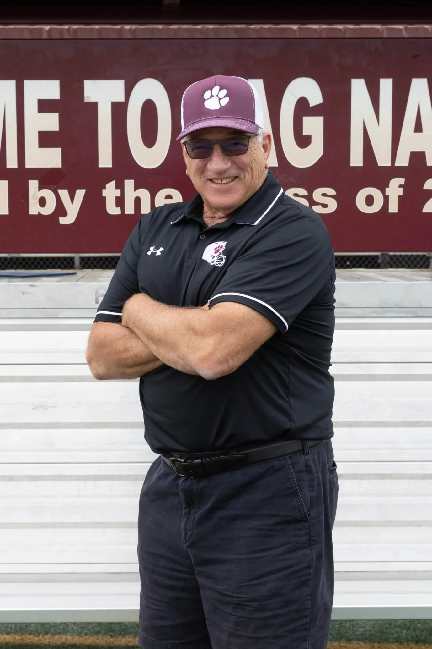 An elderly man standing on a red running track with his arms crossed, wearing a maroon polo shirt with a logo, black pants, and a baseball cap. Behind him is a landscaped area with rocks and greenery, and a sign that reads 'ON PASS' in the background.