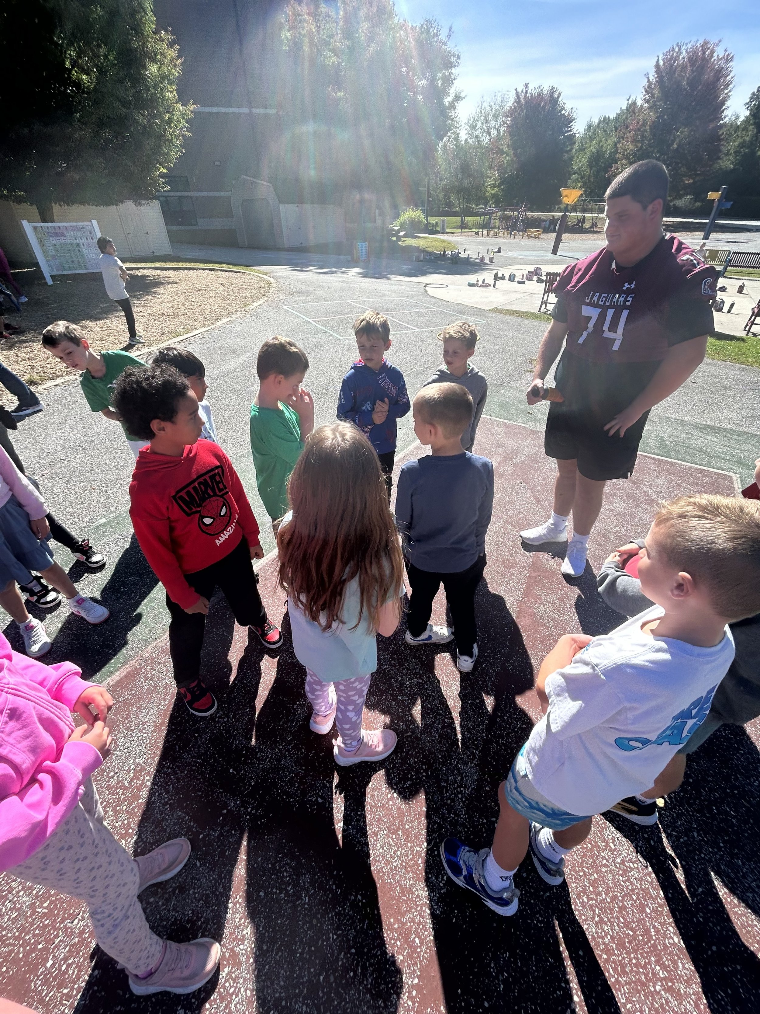 Group of children and a young man standing outdoors in a schoolyard or park, sunshine, trees, and playground equipment in the background.