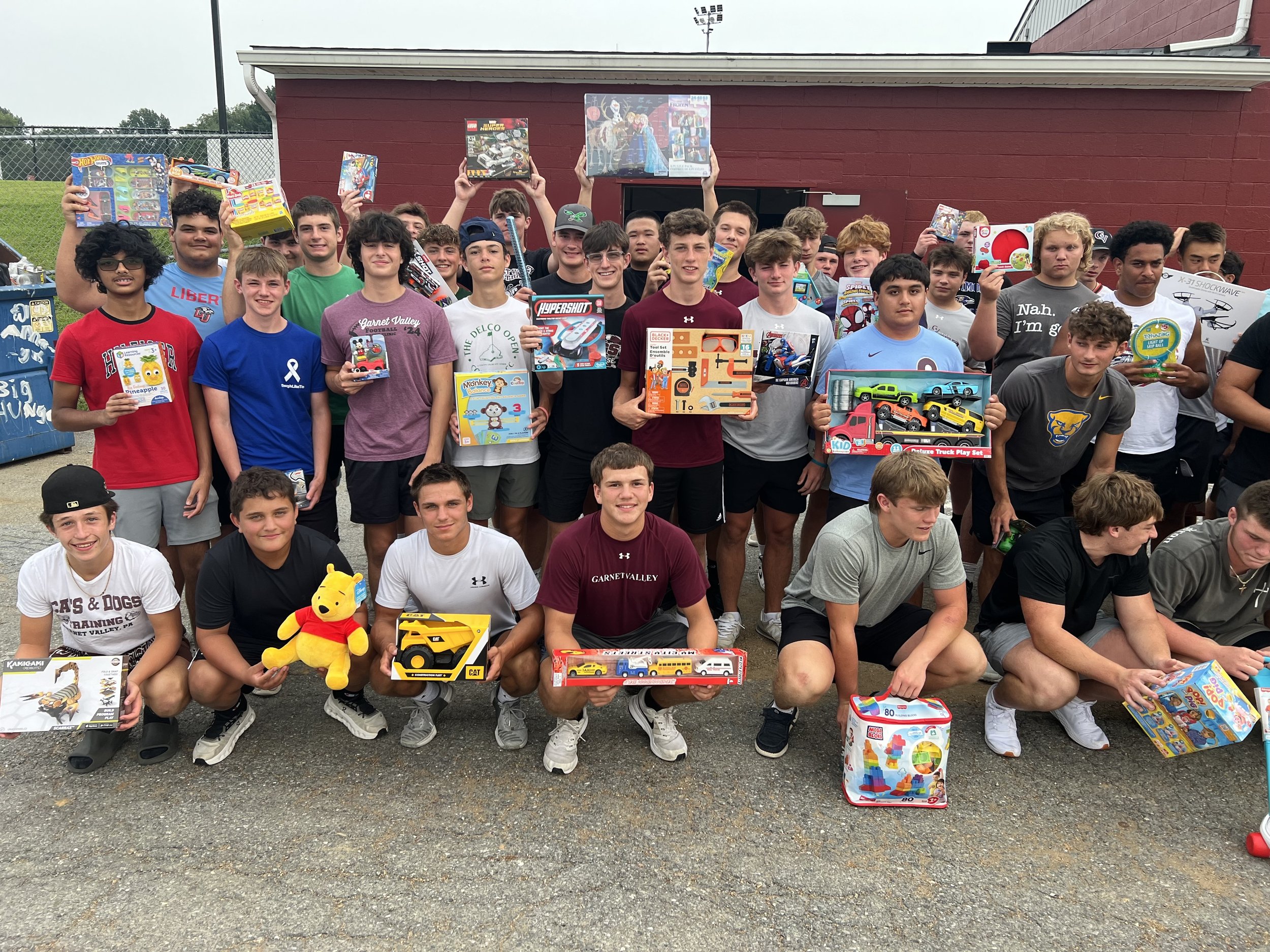 A large group of boys and teens gathered outdoors holding various toys and games, with a red building and a fence in the background.