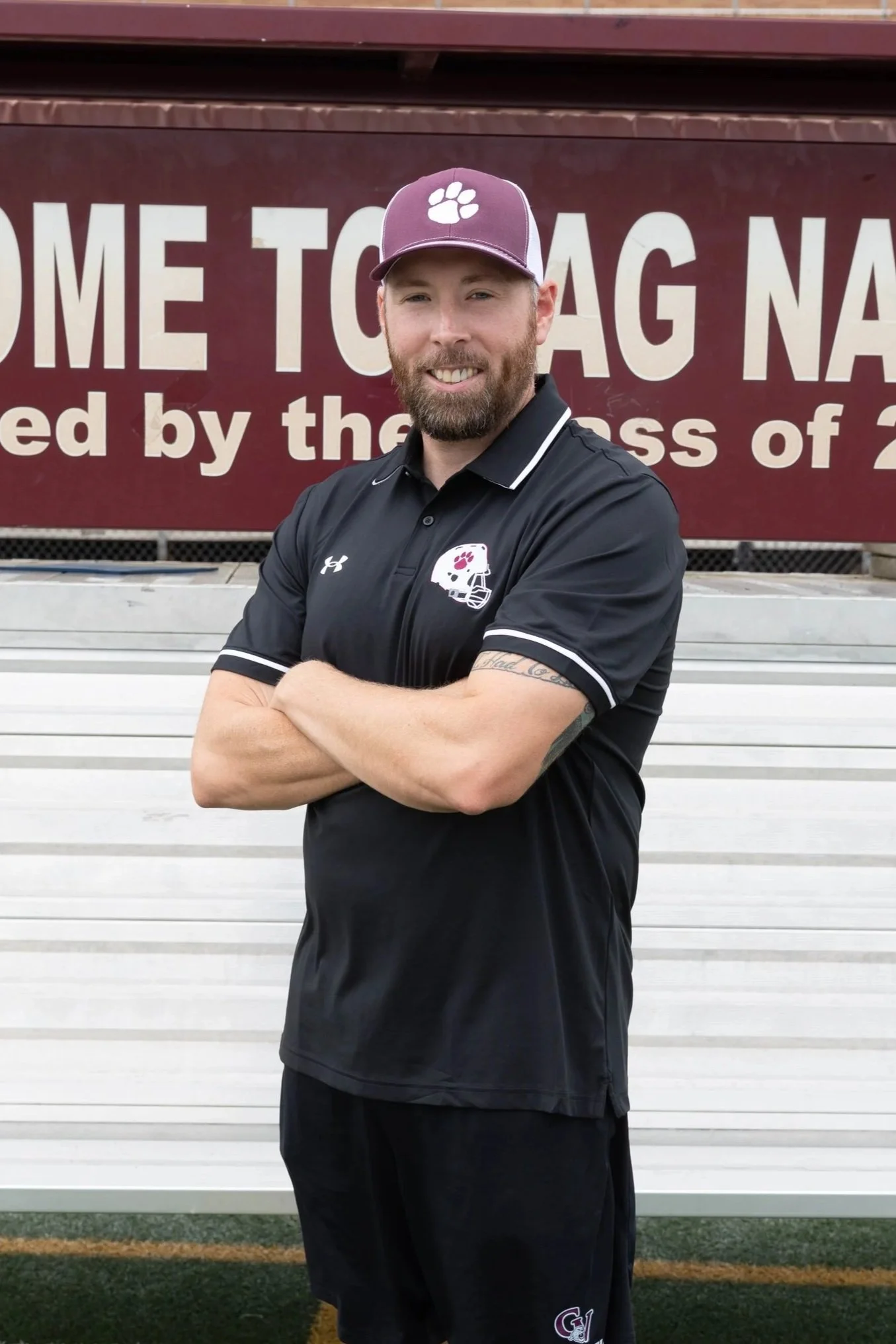 A man with a beard and sunglasses wearing a maroon polo shirt with a sports team logo, standing on a running track with arms crossed. Behind him, there is a sign that reads 'O ATHLETICS' on a hill with rocks and plants, and a cloudy sky.