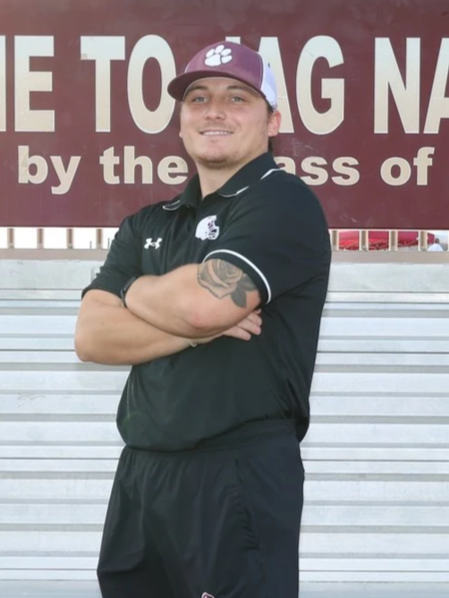 A young man with a tattoo on his left arm stands on a running track with his arms crossed, wearing a maroon athletic shirt and a gray and black cap. Behind him, there is a sign that says 'ONE NESS' and a landscaped area with rocks and plants.