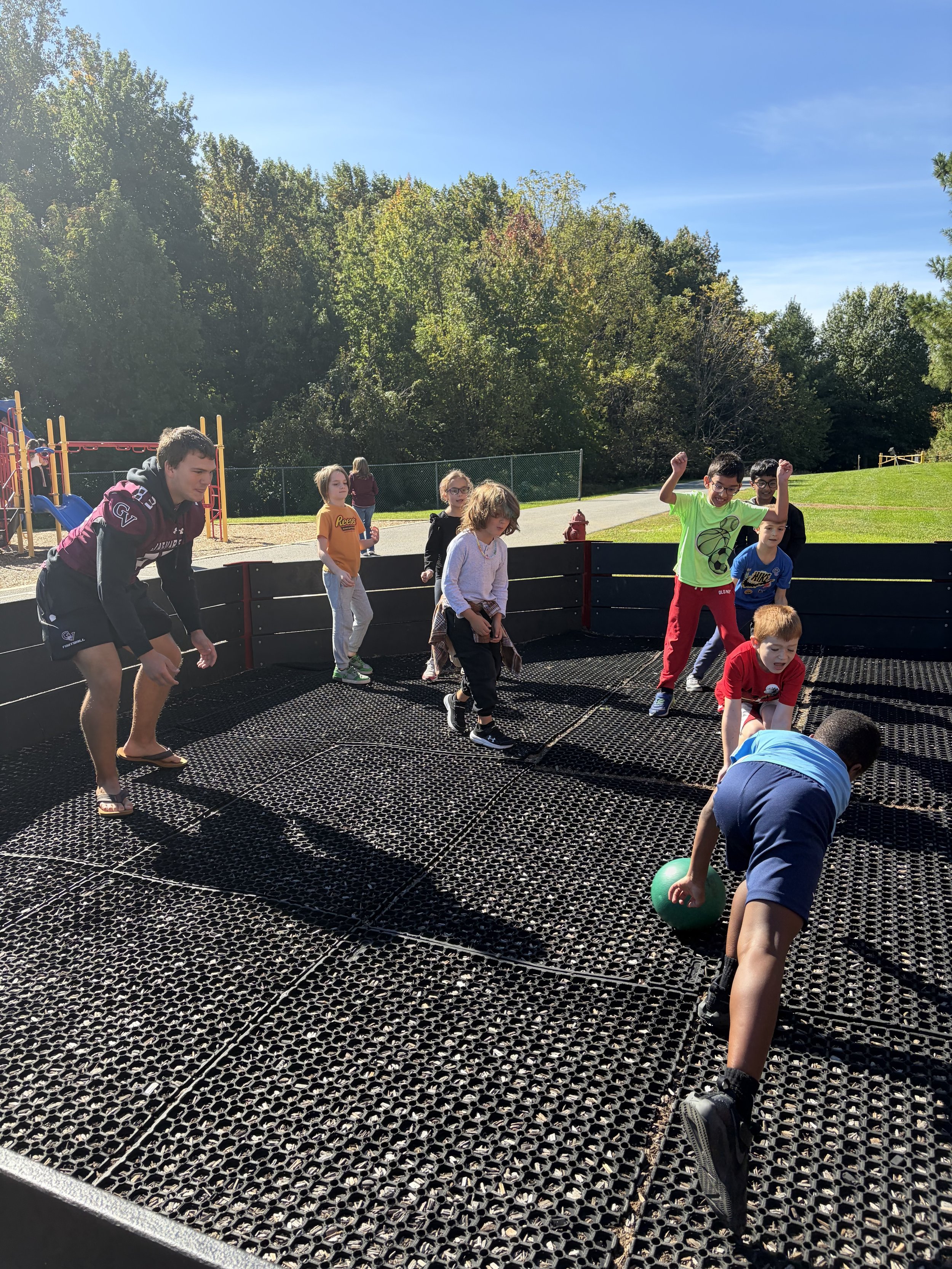 Children playing on a trampoline outdoors on a sunny day, with trees and a playground in the background.