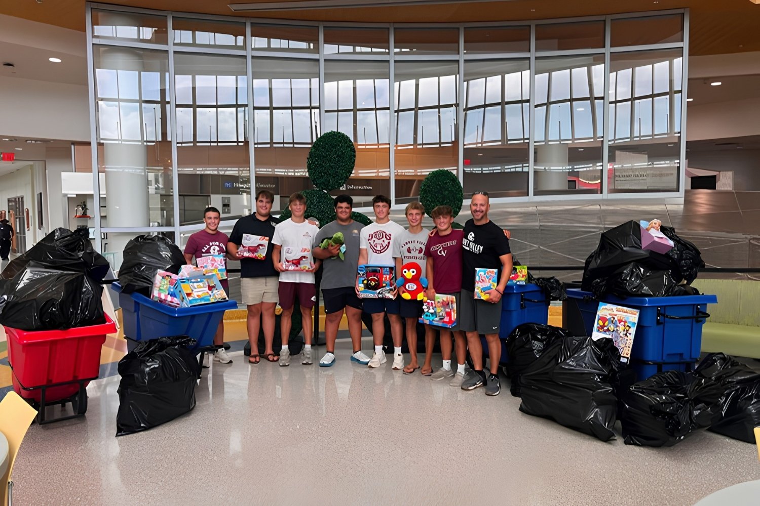 Group of eight young men standing together in an indoor public space, surrounded by large trash bags and recycling bins filled with toys and gifts.