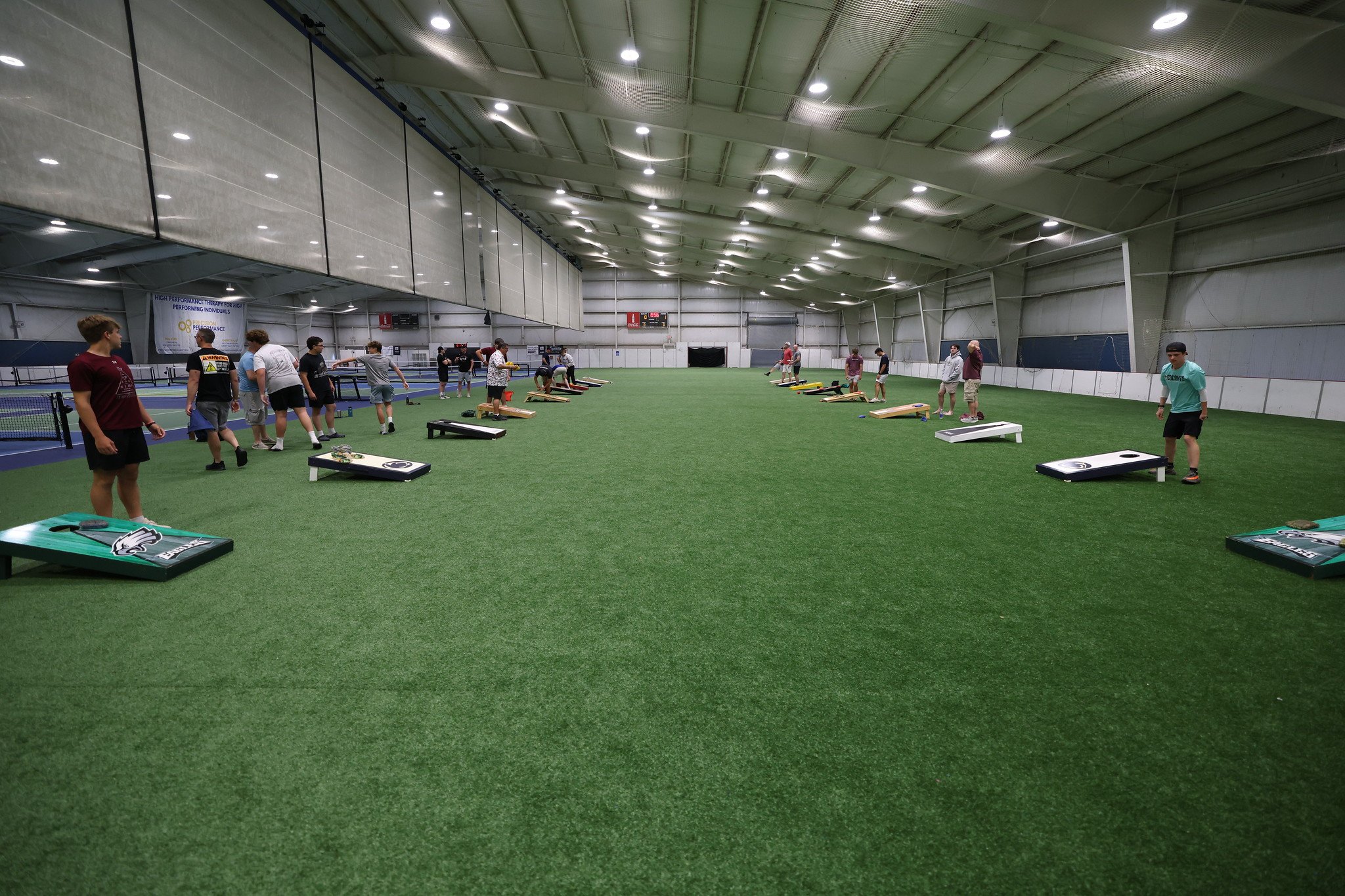 Indoor sports facility with artificial turf, people playing cornhole, and tennis courts in the background.