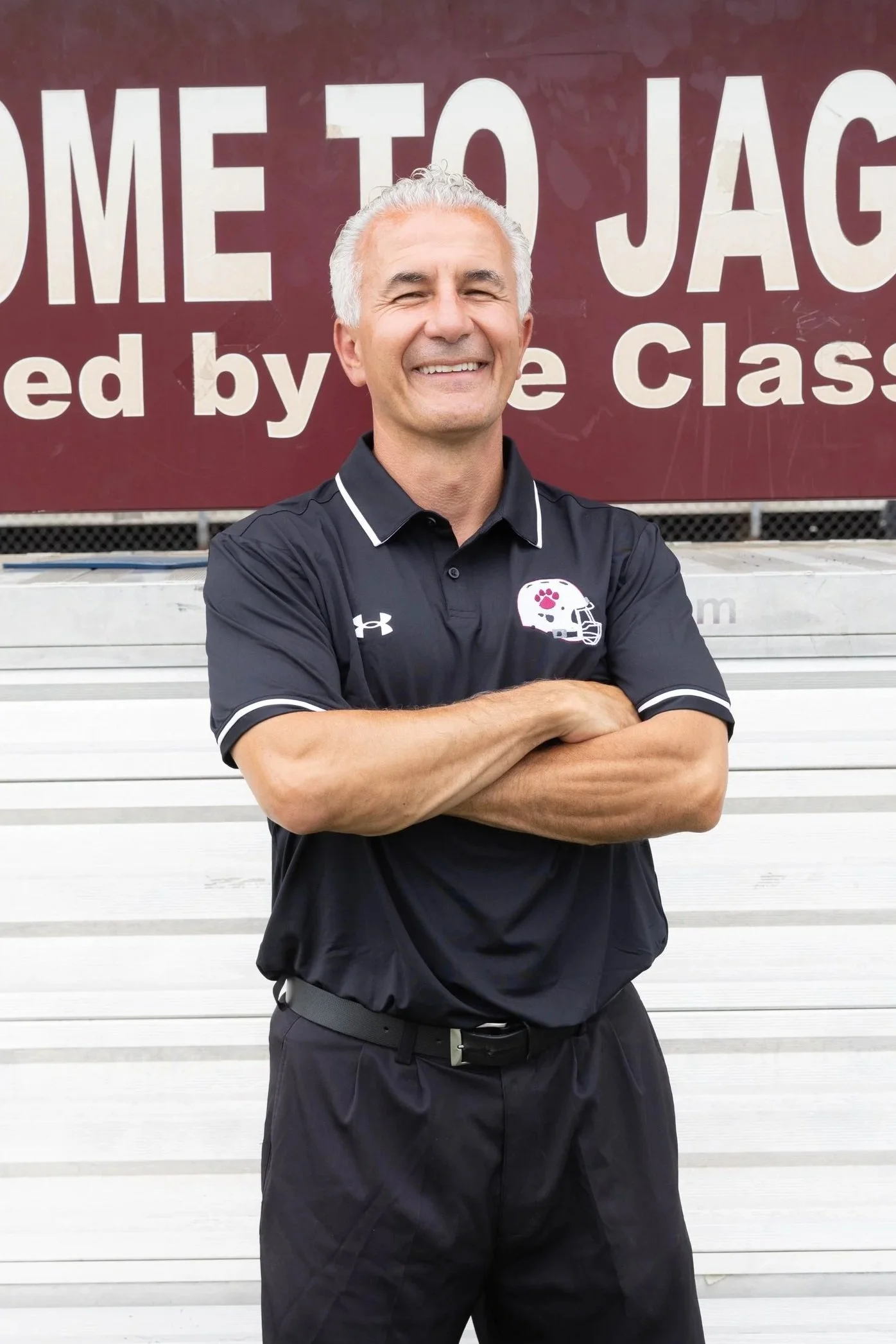 A smiling man in a maroon polo shirt and gray cap standing on a running track with arms crossed. Behind him is a large circular sign with rocks and plants, and a fence. The sign reads "Oaks" and has a logo with a bird and the letters "GU."