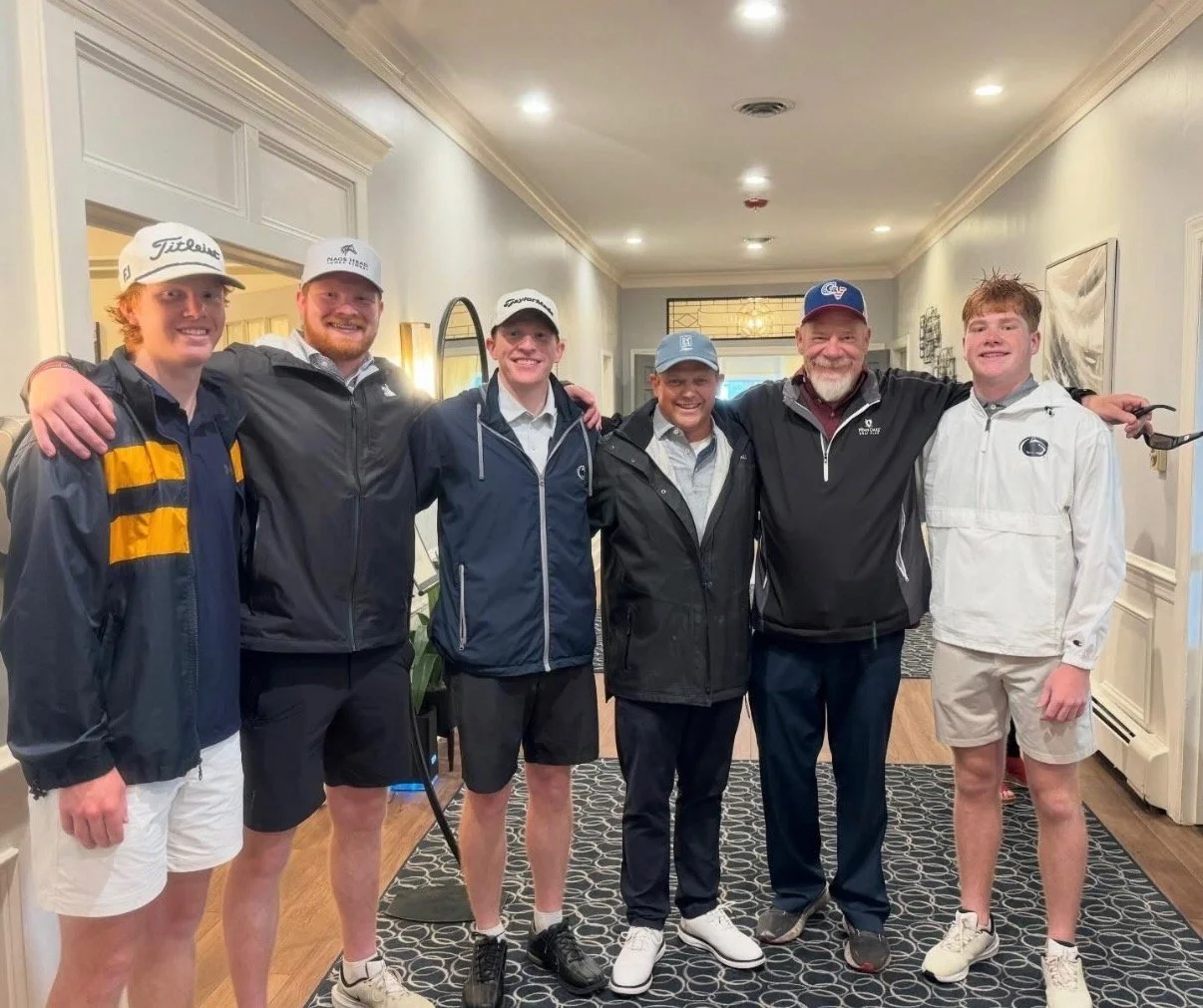 Group of six men standing together indoors, wearing golf attire and jackets, smiling for a photo.