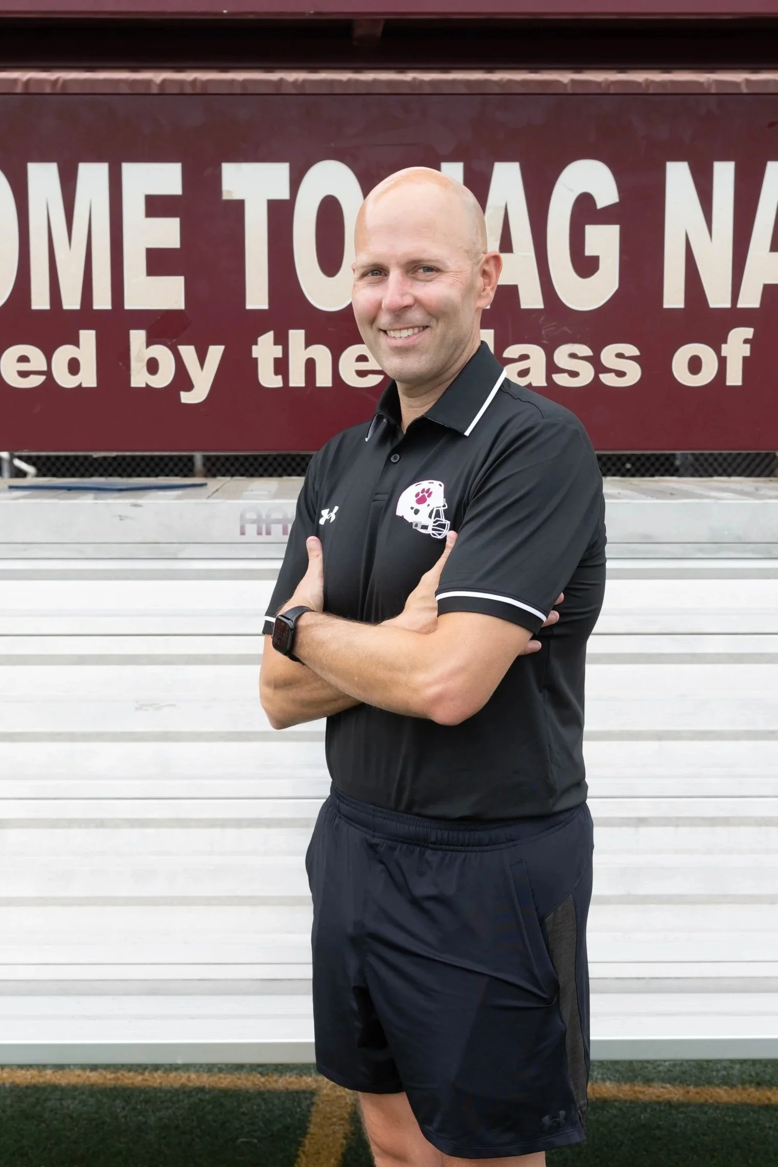 A man standing on a running track with crossed arms, wearing a maroon polo shirt with a GVSU Football logo, smiling at the camera, with a large sign in the background that says 'ON LESS' and includes a graphic of a running shoe.