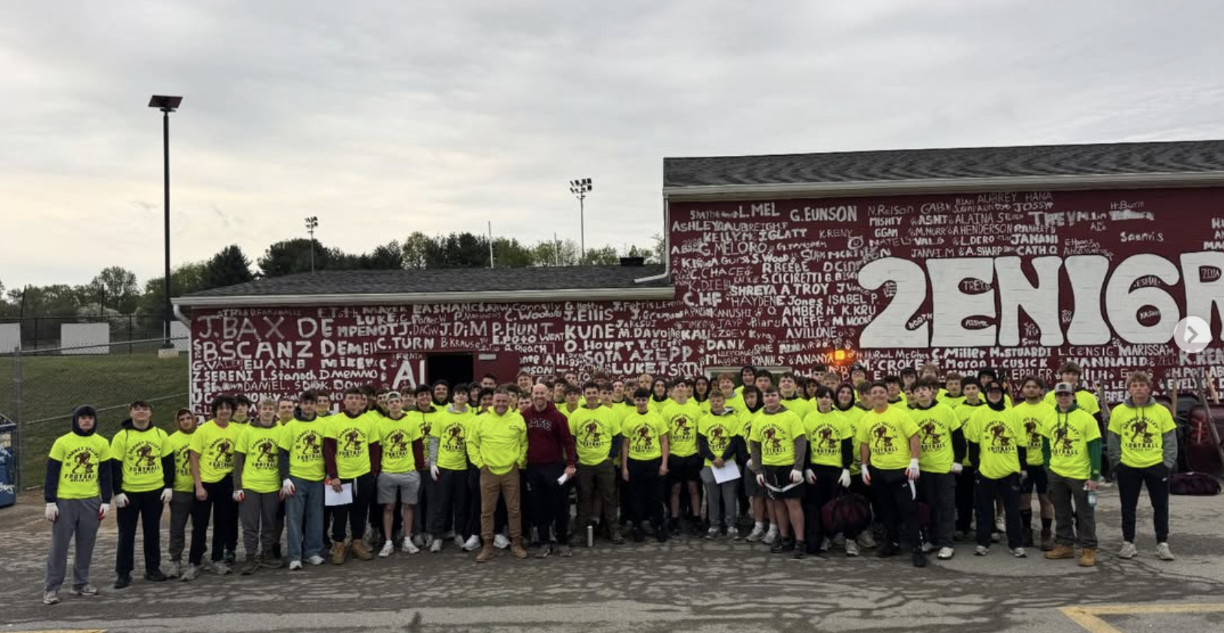 Group of young people wearing bright yellow T-shirts and gloves, standing together outdoors in front of a large red wall with white graffiti and a white 'SPORTS' text, participating in a community event or team activity.