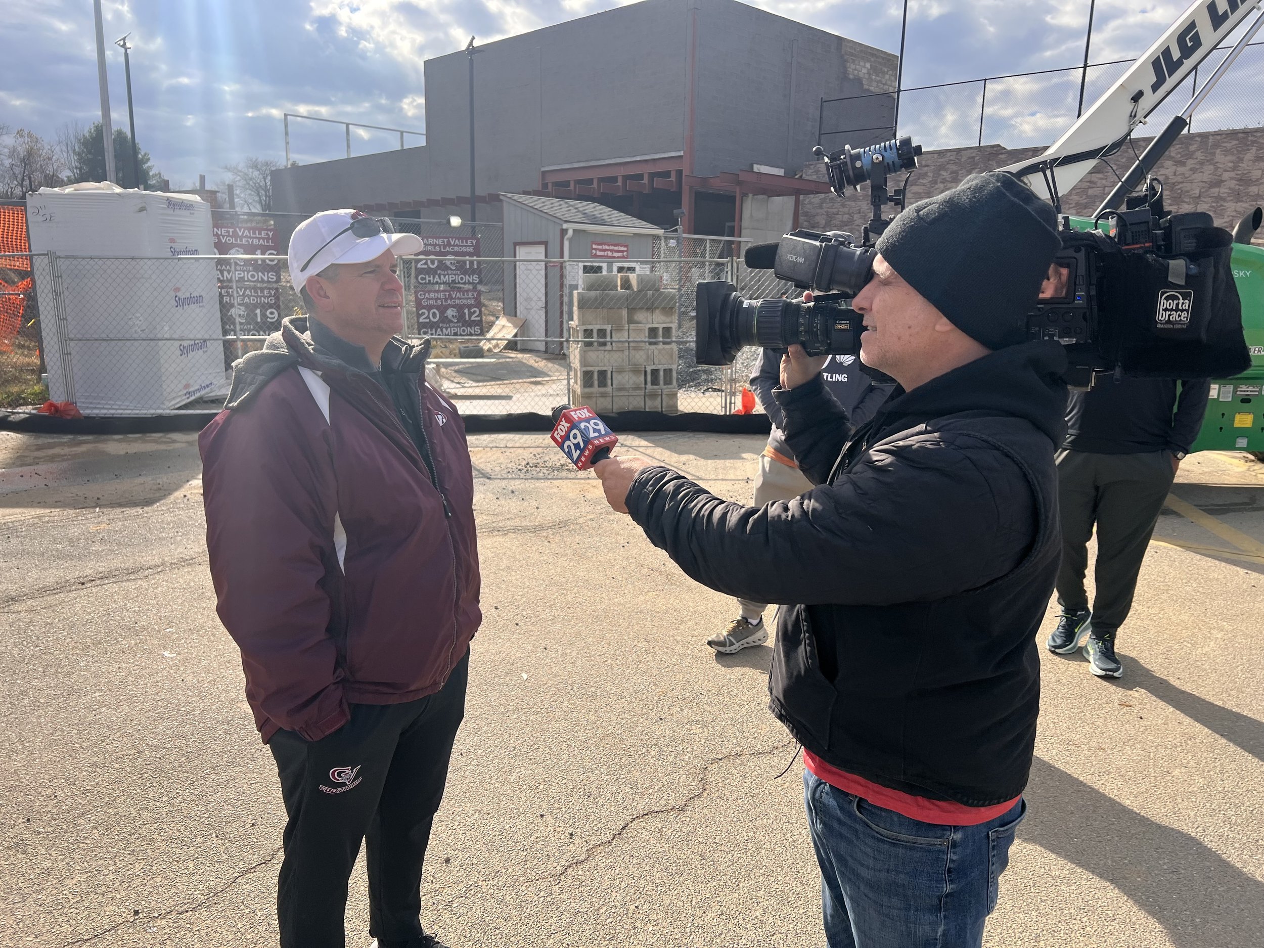A man in a maroon jacket and white cap being interviewed by a man holding a FOX 29 microphone. A camera operator records the interview with a professional video camera. They are outdoors near a construction site with building materials and a chain-link fence in the background.