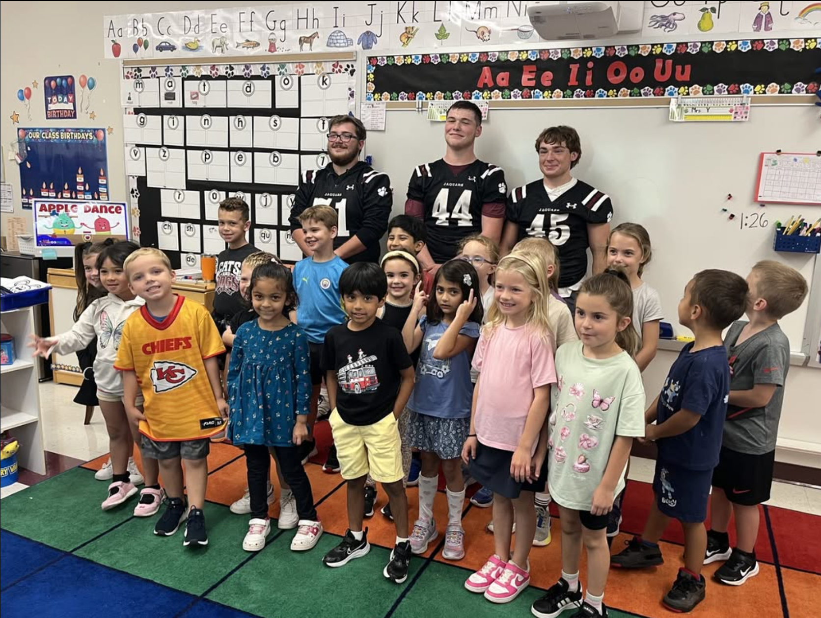 Group of young children and three teenage boys standing in a classroom, smiling at the camera. The children are of diverse ethnicities, dressed in casual clothes, some with patterns or sports team logos. The teachers are wearing football jerseys. The classroom has educational posters, including the alphabet on the wall, class birthday calendar, and various decorations.