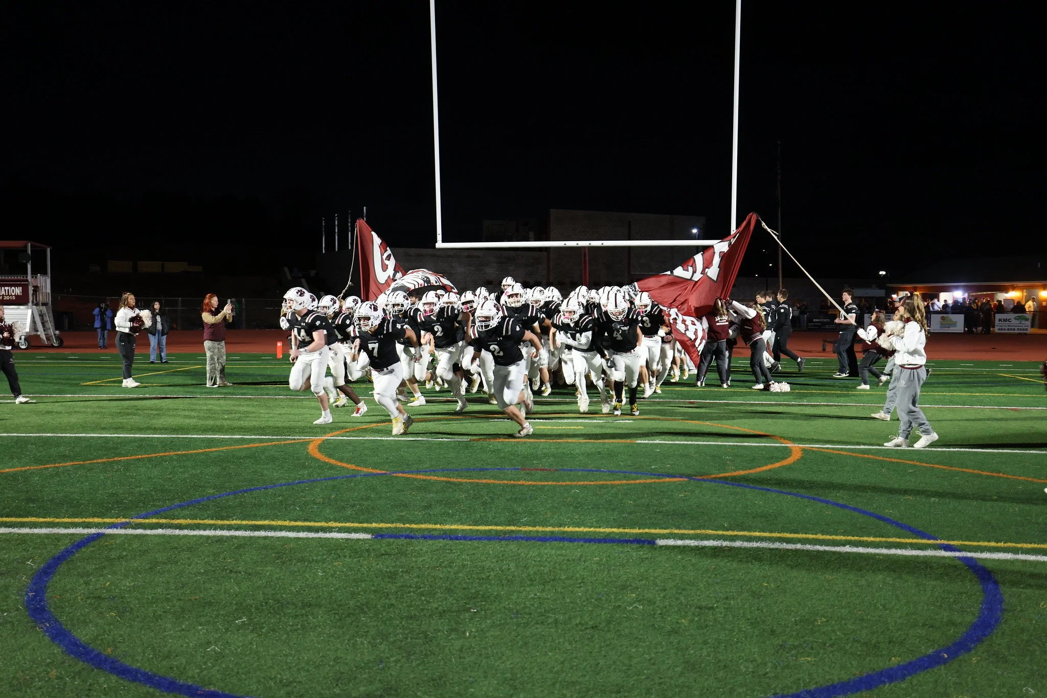 High school football team running onto the field through a large banner held by cheerleaders during a night game at a stadium.
