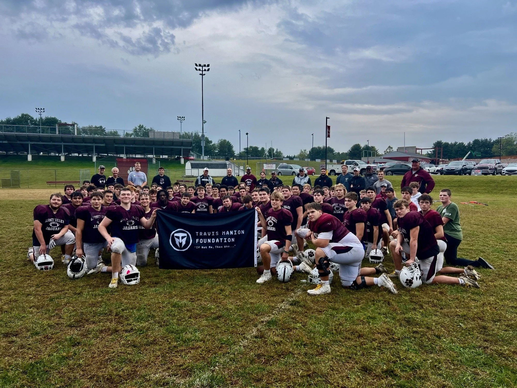 A large group of football players and coaches pose for a team photo on a football field. The players are wearing maroon jerseys and white pants, with some holding helmets on the ground. The coaches stand behind the players. The team is holding a blue banner that reads 'Travis Manion Foundation.' The background shows a grassy field, stadium seating, and parked cars under a partly cloudy sky.