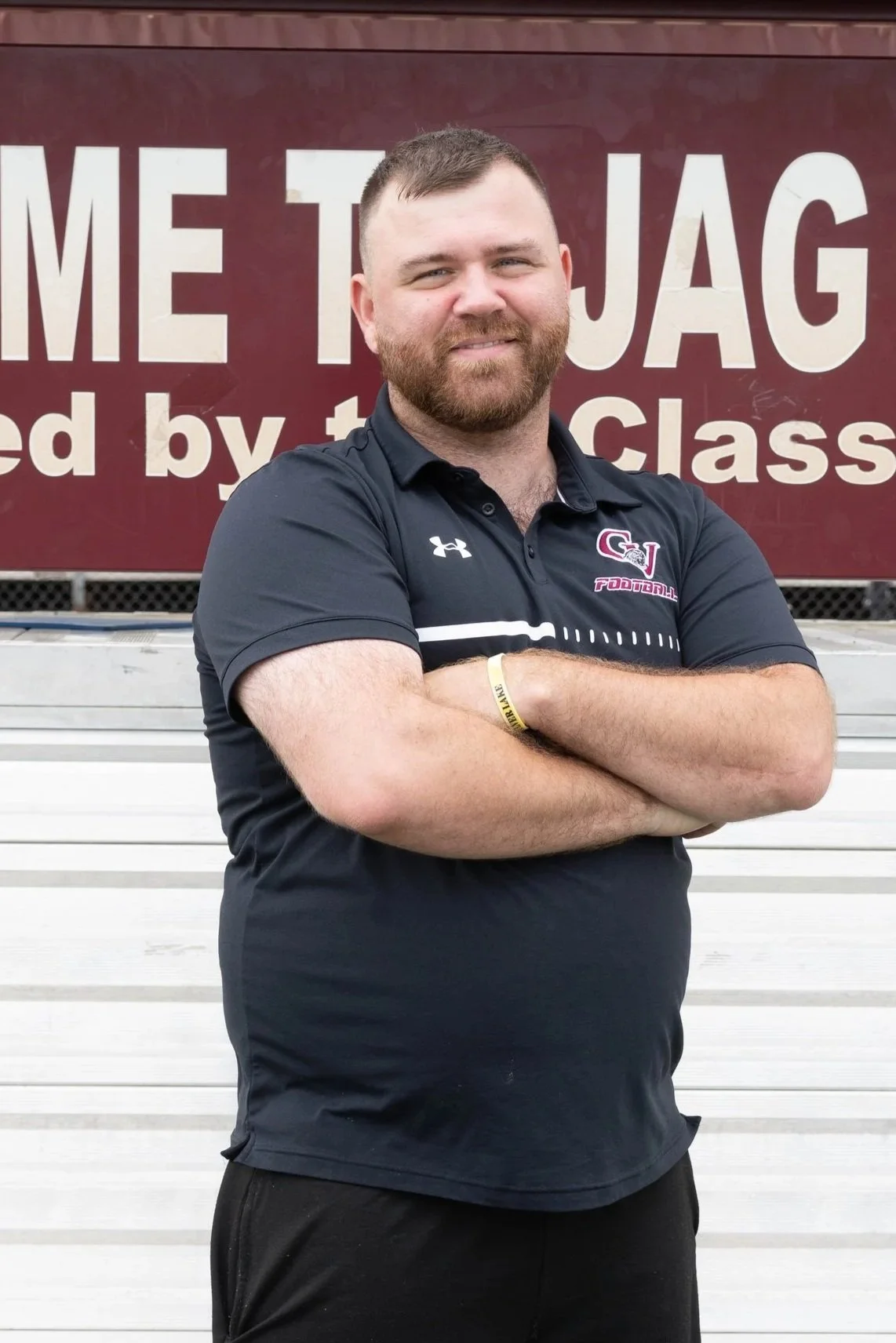 A man with crossed arms standing on a running track at a football field, with a large emblem and a sign in the background.