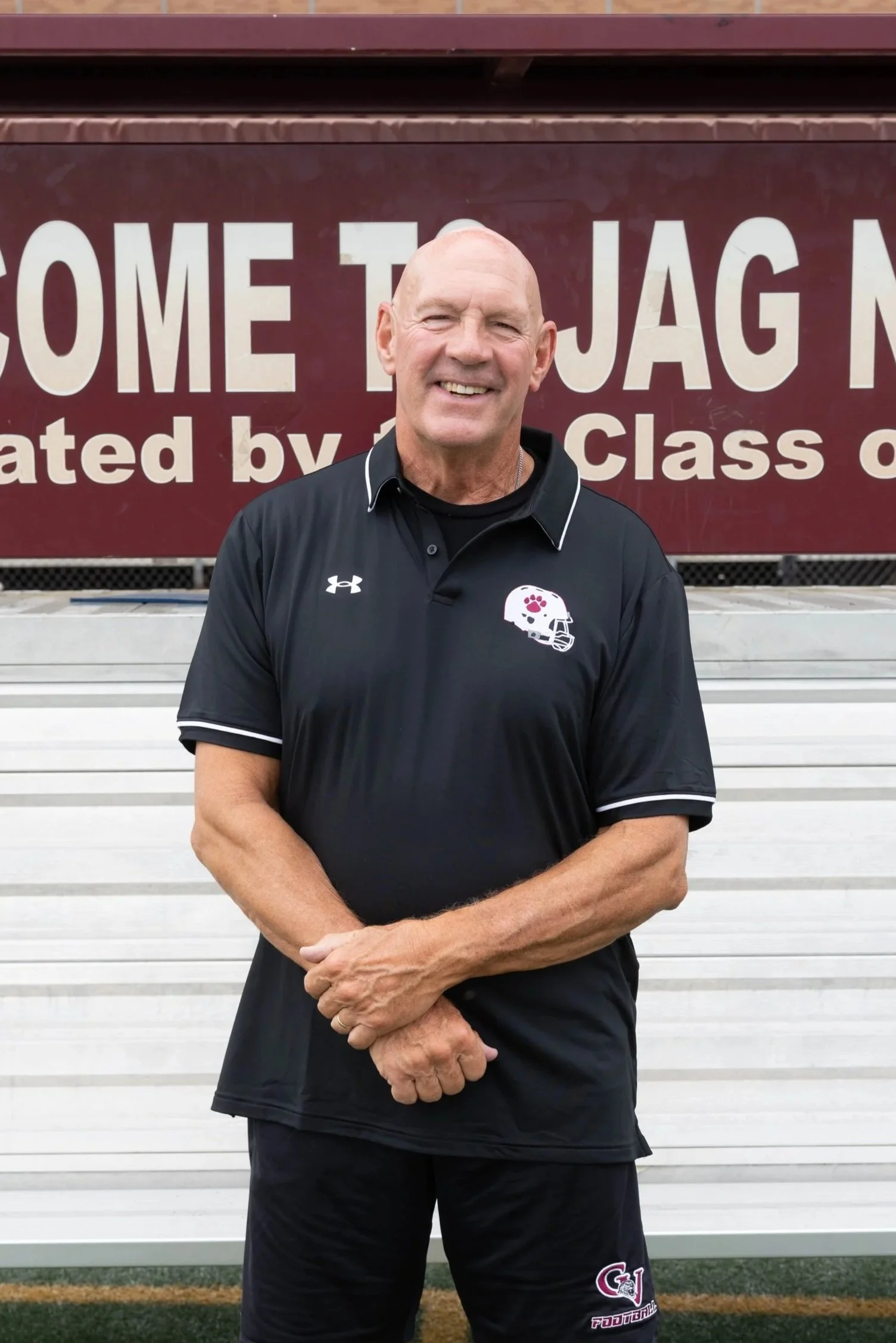 A smiling man in a maroon sports shirt stands on a running track with folded arms. In the background, there is a circular decorative area with rocks, greenery, and a sign that reads 'ONE GOAL'.