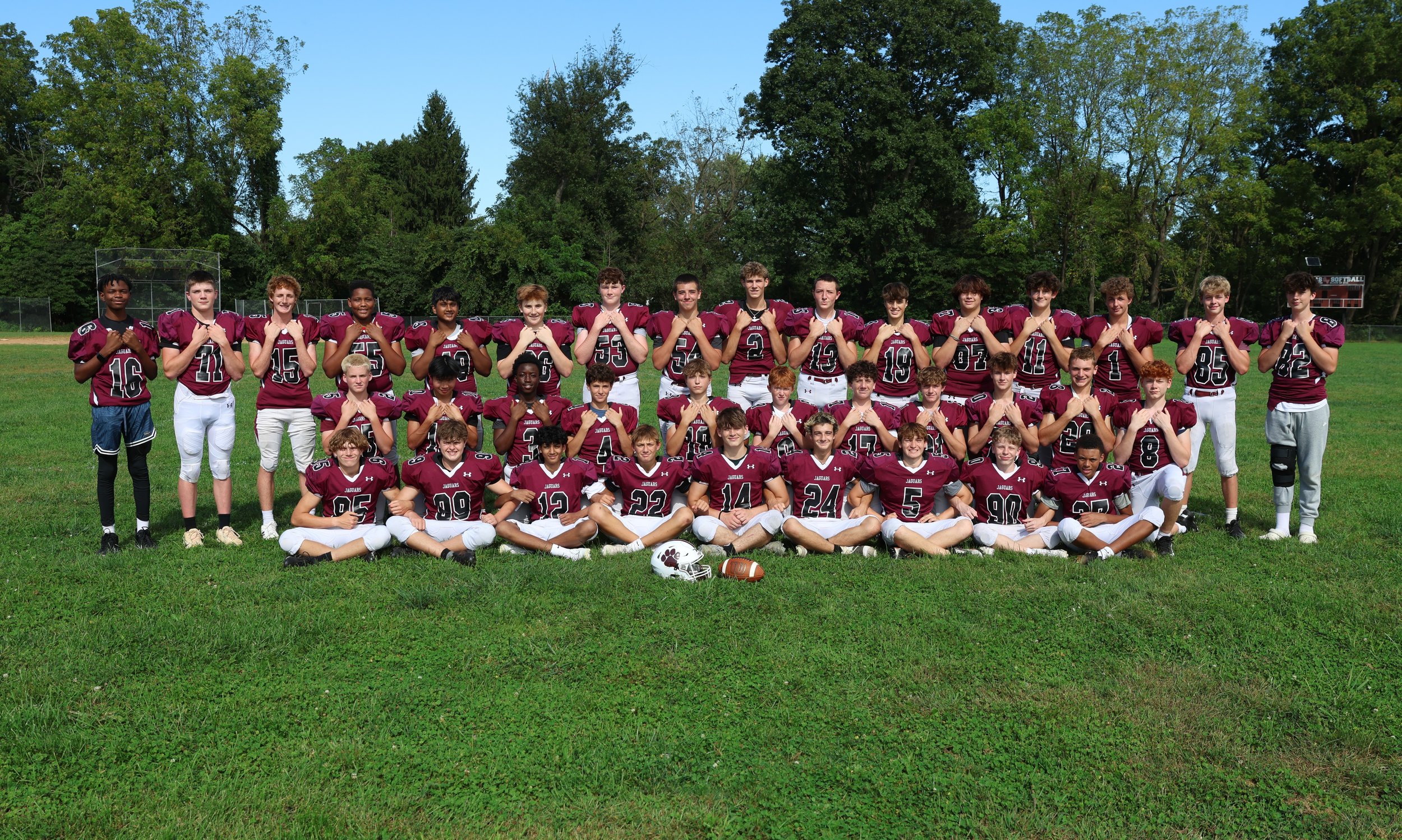 Group of football players in maroon jerseys posing on a grassy field with trees in the background.