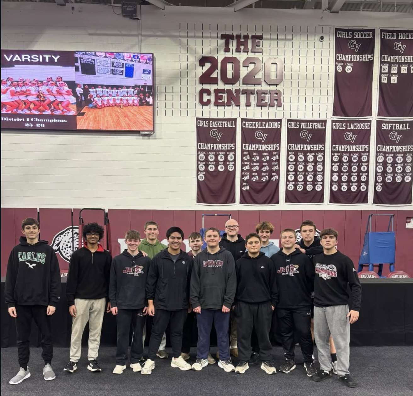 A group of 13 young male athletes standing inside a gymnasium, posing for a photo. The gymnasium has banners on the wall indicating various sports championships and a large screen displaying a volleyball team. The team members are dressed in casual sportswear, with some wearing hoodies, sweatshirts, and sweatpants.