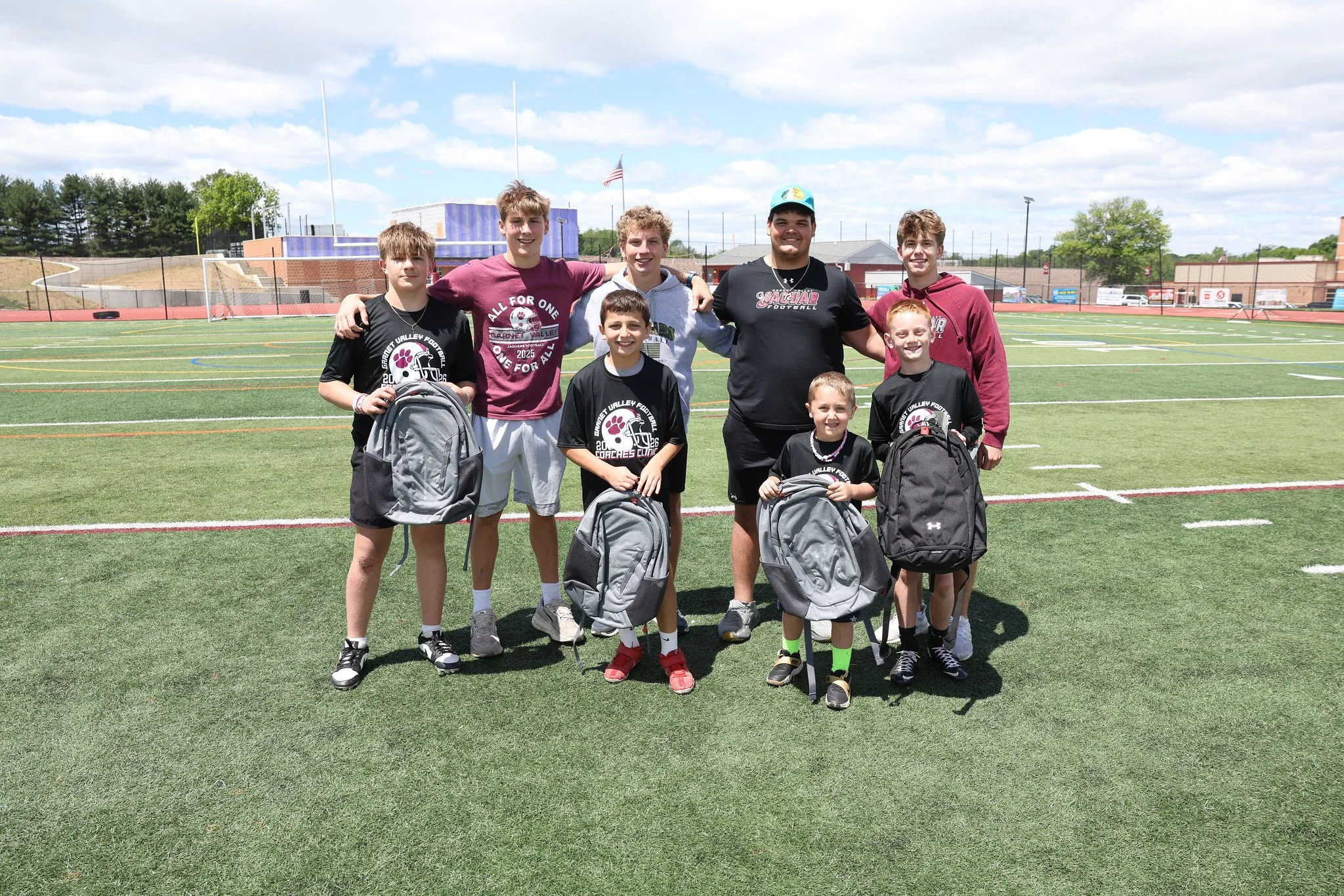 Group of young boys and a coach standing on a football field holding backpacks, smiling for a photo on a sunny day.