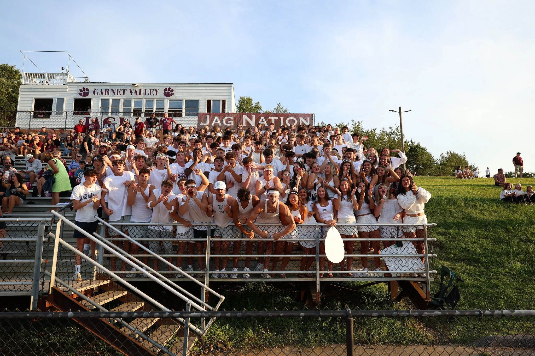 A large crowd of students sitting on stadium bleachers at Garnet Valley High School, with some standing and talking, at night time.