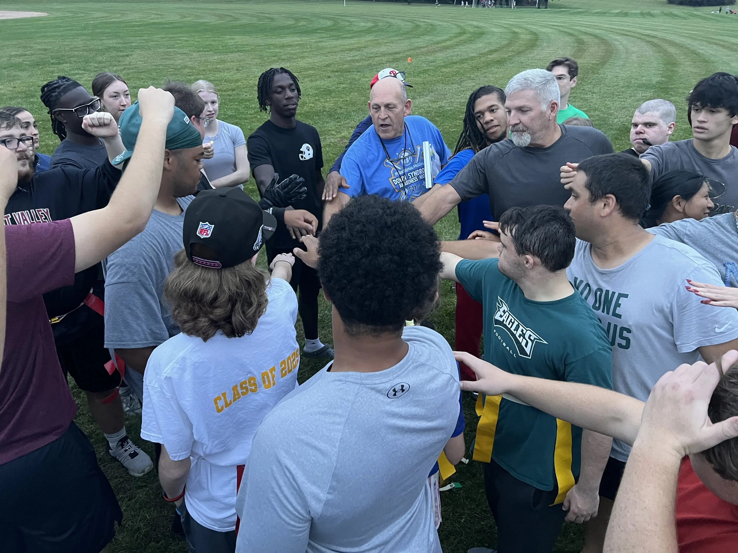 A group of people, including athletes and coaches, are gathered in a circle on a grassy field, participating in a team huddle or meeting.