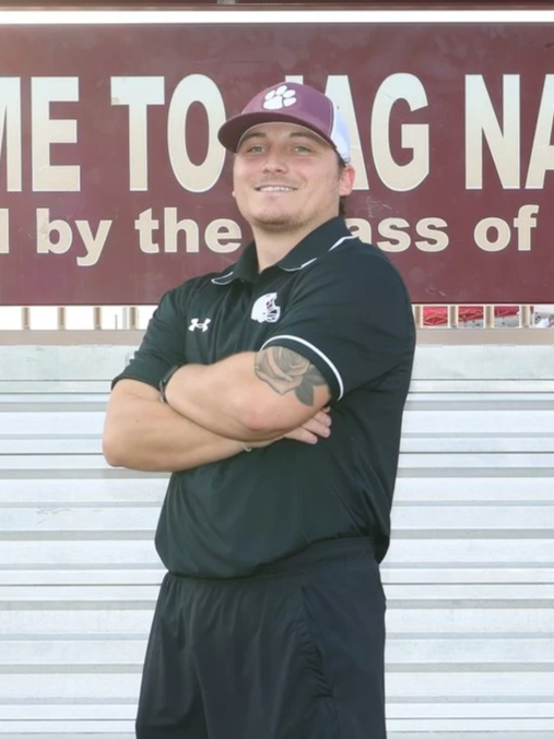 A young man with a tattoo on his left arm stands on a running track with his arms crossed, wearing a maroon athletic shirt and a gray and black cap. Behind him, there is a sign that says 'ONE NESS' and a landscaped area with rocks and plants.