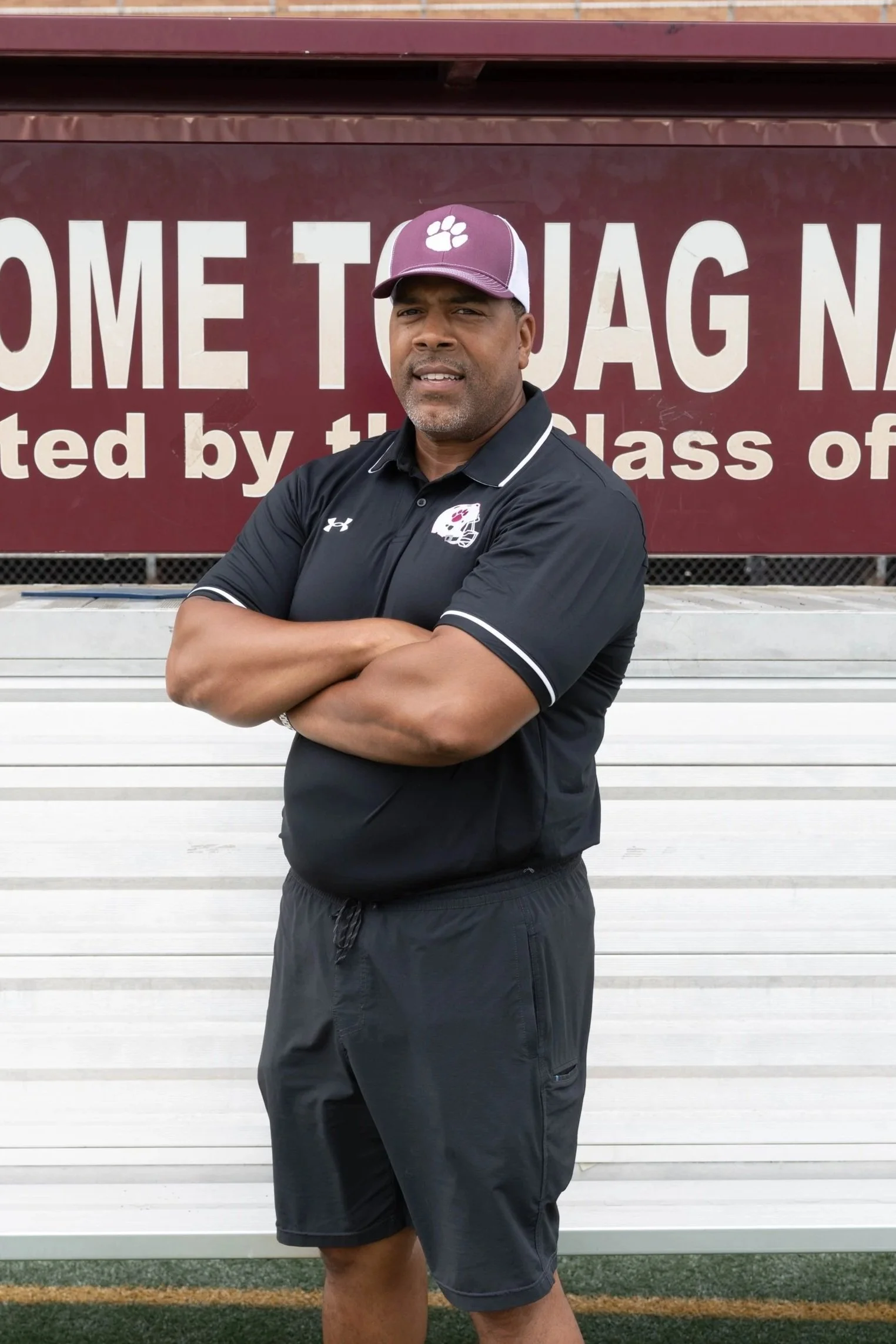 Man standing on a red running track with arms crossed, wearing a maroon polo shirt, sunglasses, and a black and gray baseball cap. Behind him is a decorative hill with rocks and a sign that says 'ON'. The sky is cloudy.