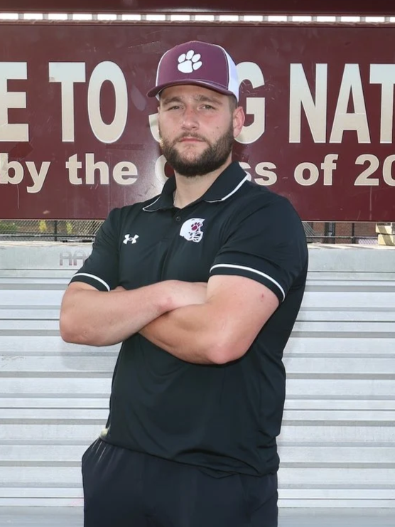 Young man standing with arms crossed on a running track in front of a hill and a sign that says 'ONE'. He is wearing a maroon football shirt and a gray cap with a logo.