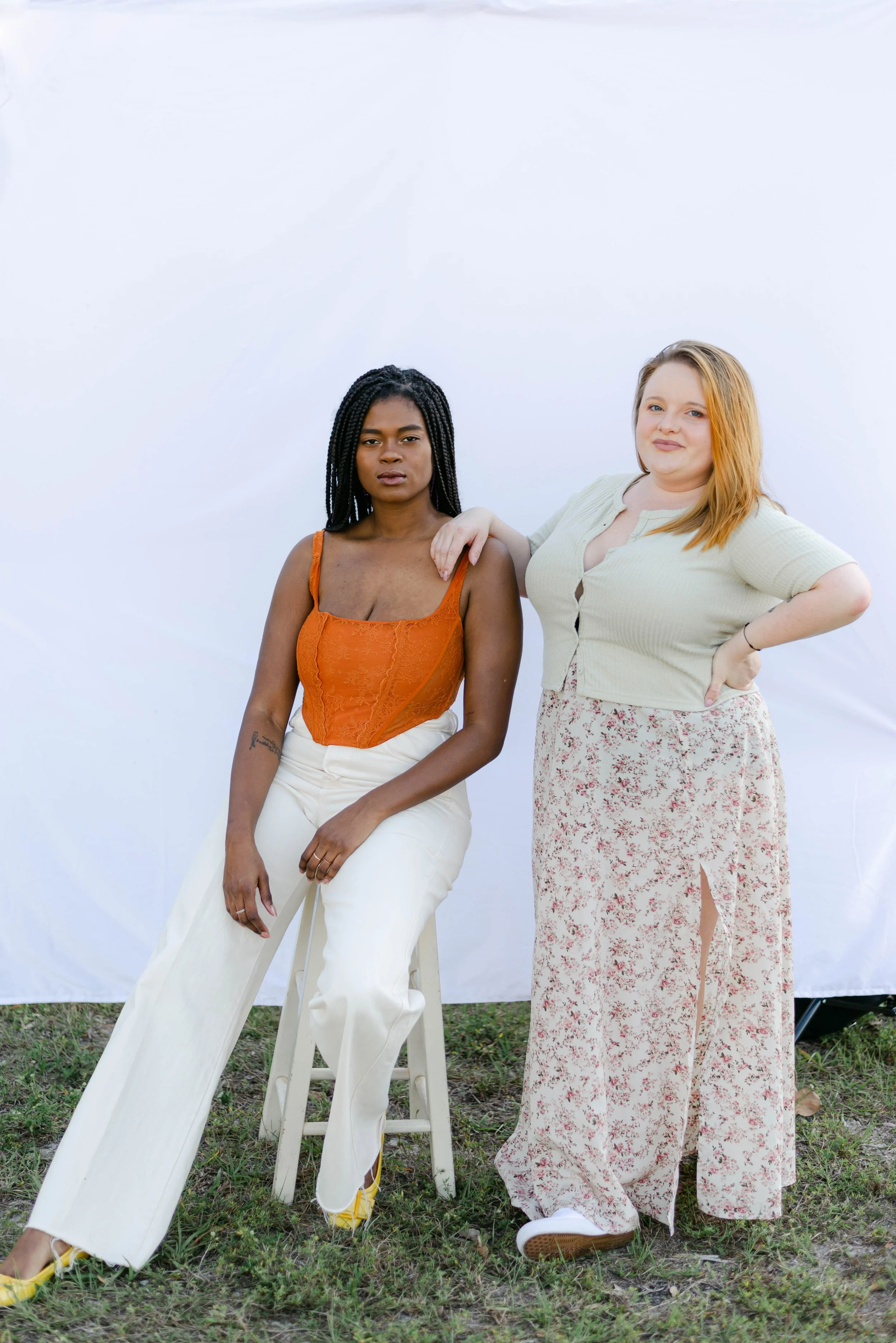 Two women standing in front of a white background outdoors, one seated on a stool and the other standing with her hand on the seated woman's shoulder, both wearing casual outfits.