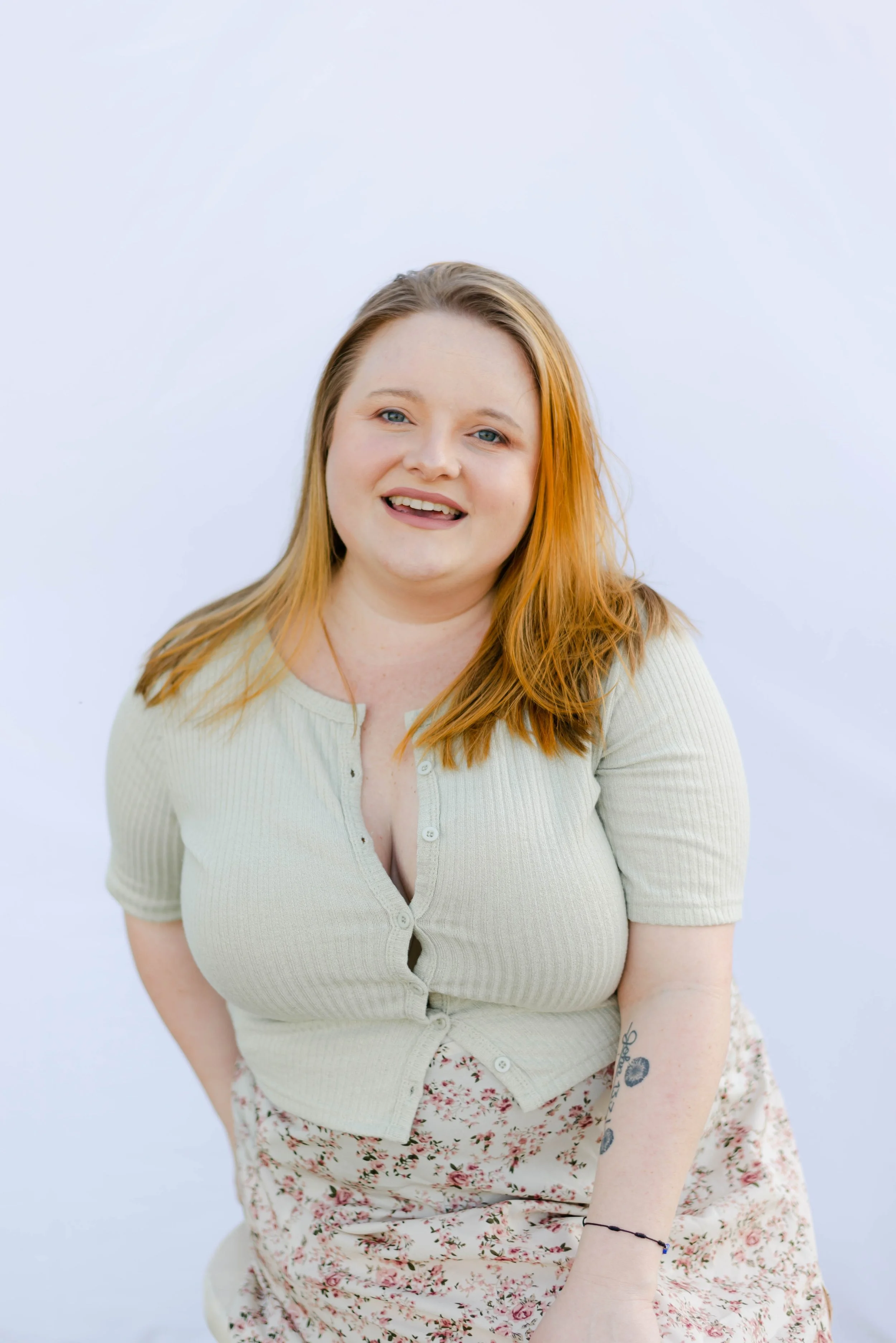 One of the founders of Post Glow Studio, smiling in front of a white backdrop, wearing a light green top and floral skirt with a visible forearm tattoo.