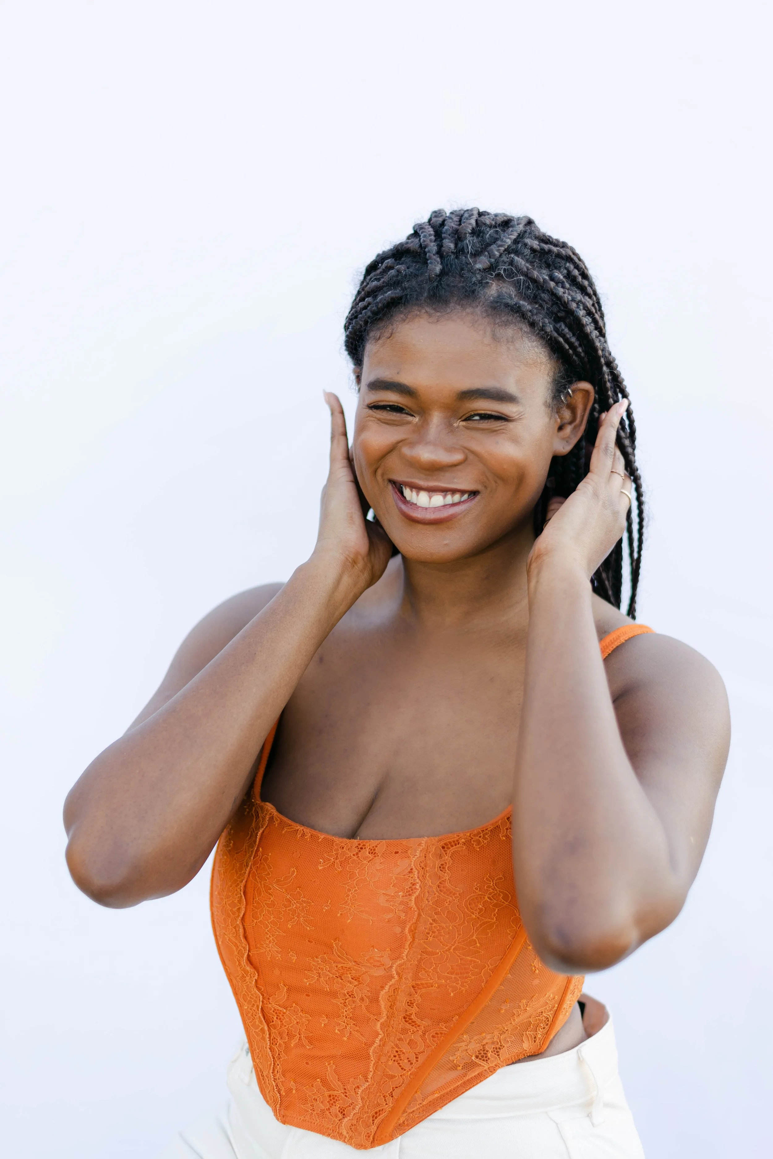 One of the founders of Post Glow Studio smiling brightly in front of a white backdrop, wearing an orange lace top and white pants, with hands playfully touching her face.