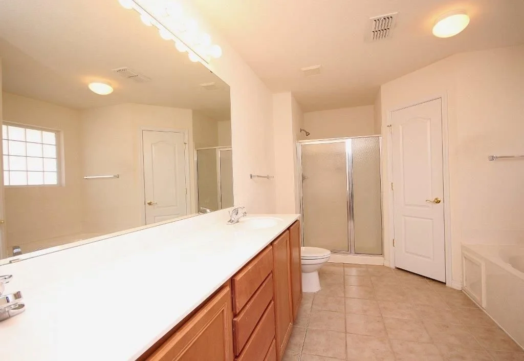 Bathroom with a large mirror, double sink vanity, toilet, shower with frosted glass door, and beige tiled floor.