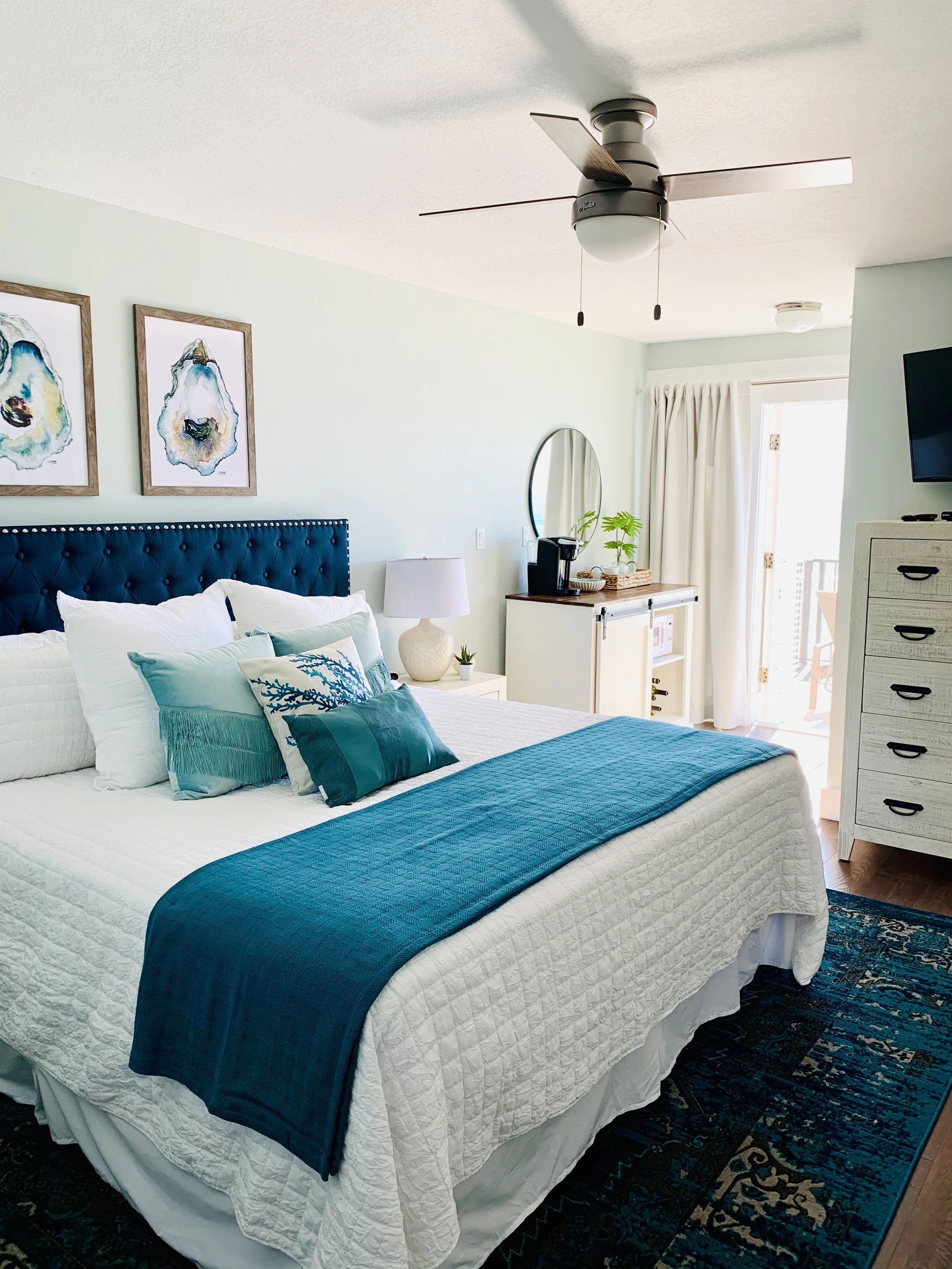 A well-lit bedroom with a white bed, blue accent pillows, and a blue bedspread. There is a ceiling fan with lights, artwork above the bed, a white nightstand with a lamp, and a dresser with a decorative mirror and a plant. A sliding glass door with w
