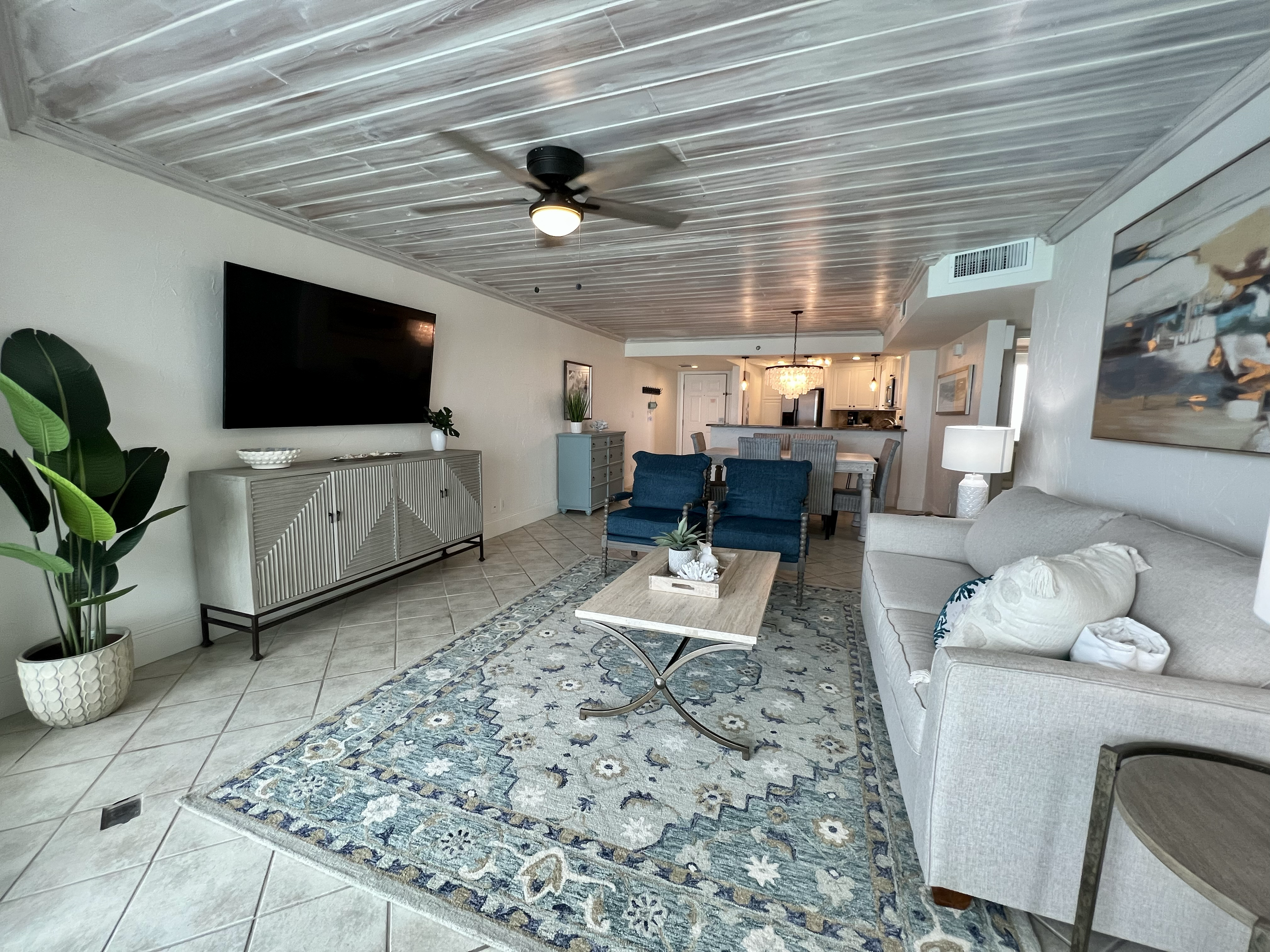 Living room with white sofa, blue armchairs, a wooden coffee table, a patterned rug, a flat-screen TV on a console, potted plants, a painting, and a view into the kitchen area.