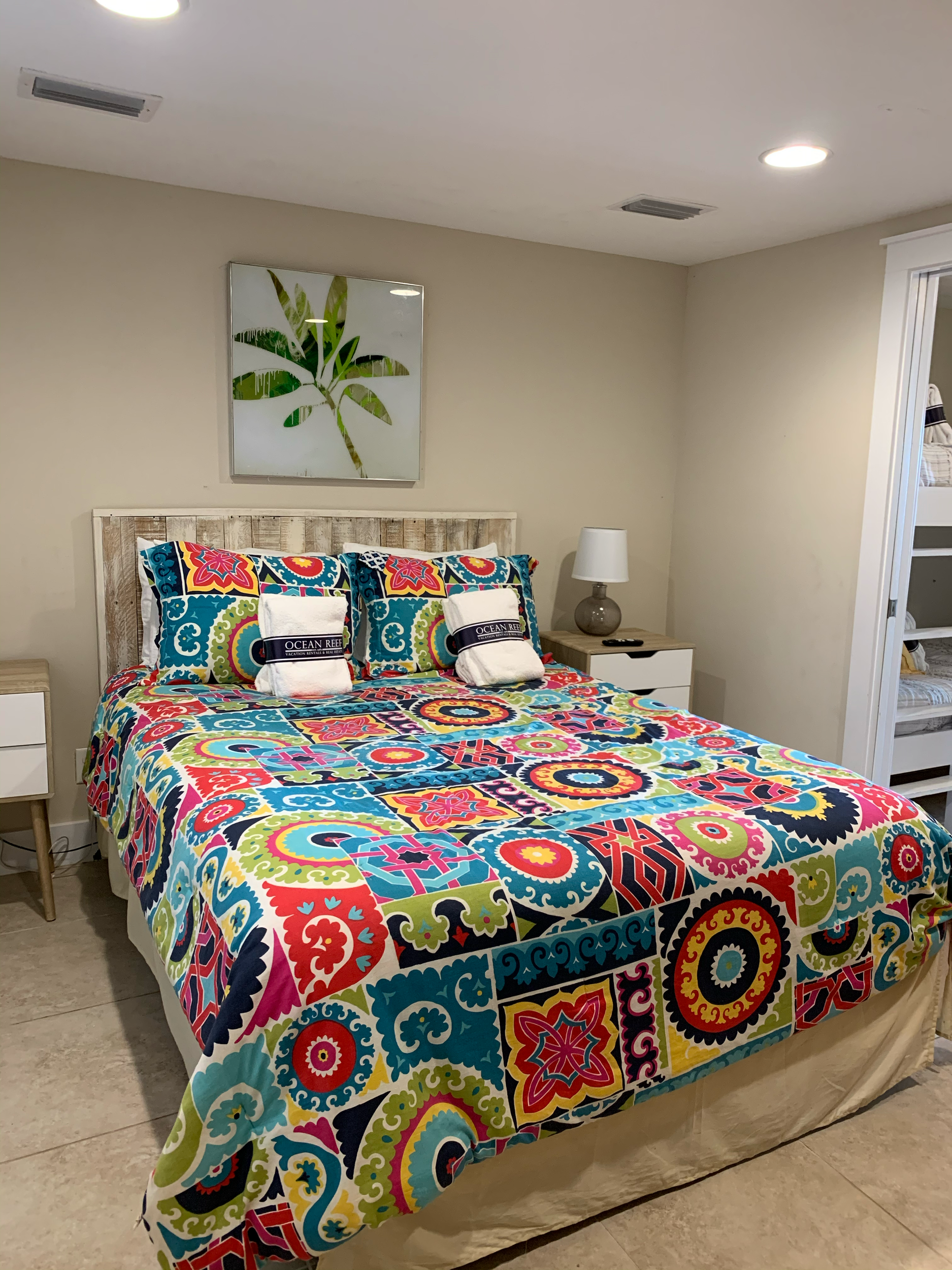 Bedroom with a bed covered in a colorful quilt, two pillows, and a towel with the Ocean Reef logo. There is a nightstand with a lamp, and a framed leaf artwork above the bed.